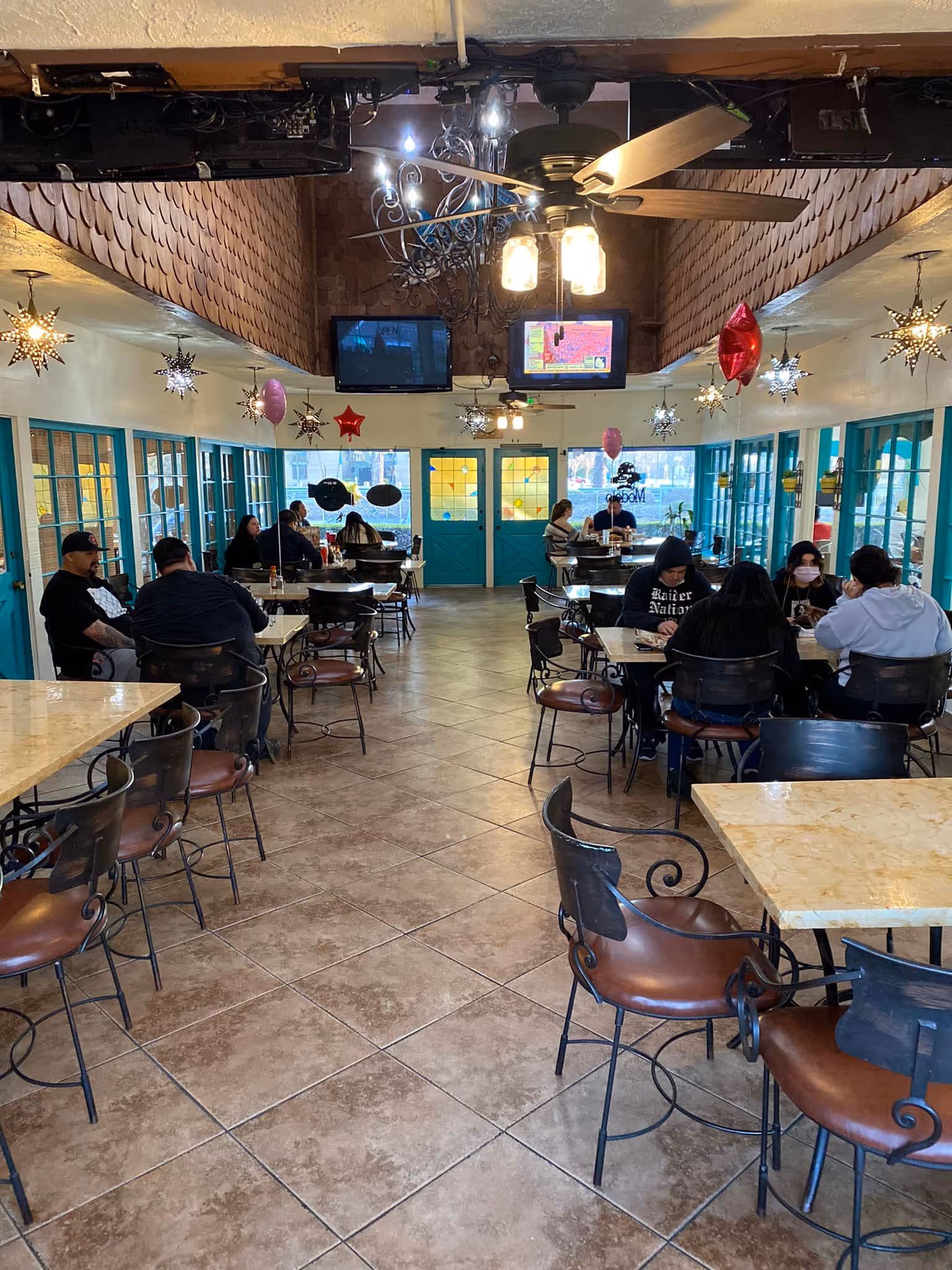 Interior of a restaurant with tiled floor, turquoise doors and windows, star-shaped hanging lights, balloon decorations, and people seated at tables.