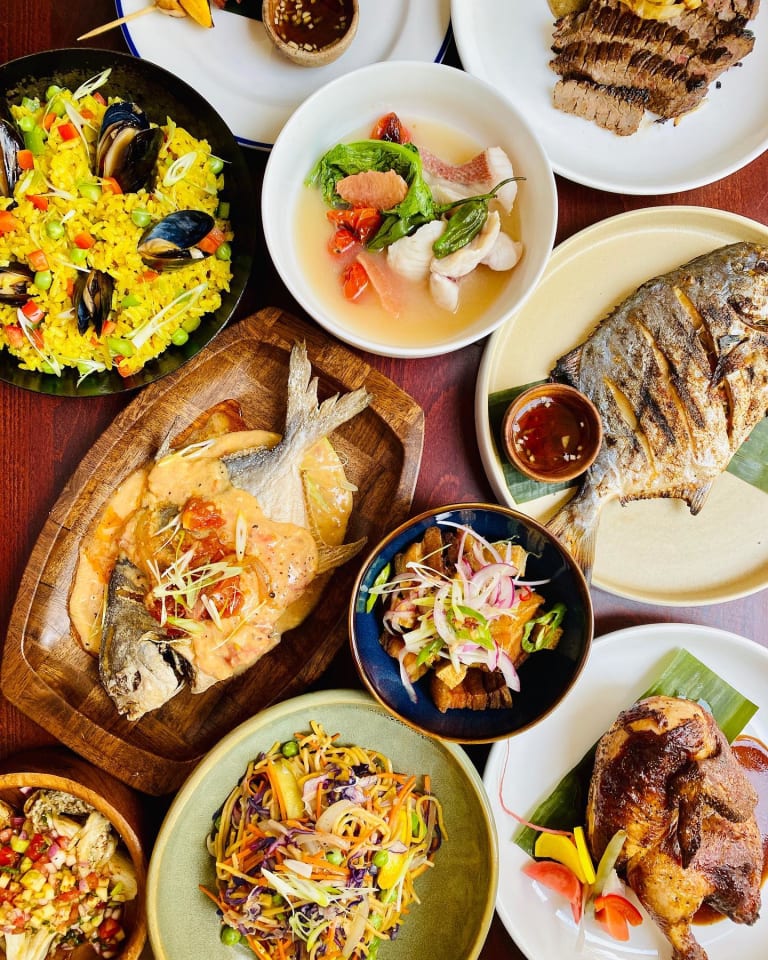 A family-style spread of Musang dishes including paella with mussels, whole grilled fish, lechon kawali, pancit, and rotisserie chicken on a wooden table