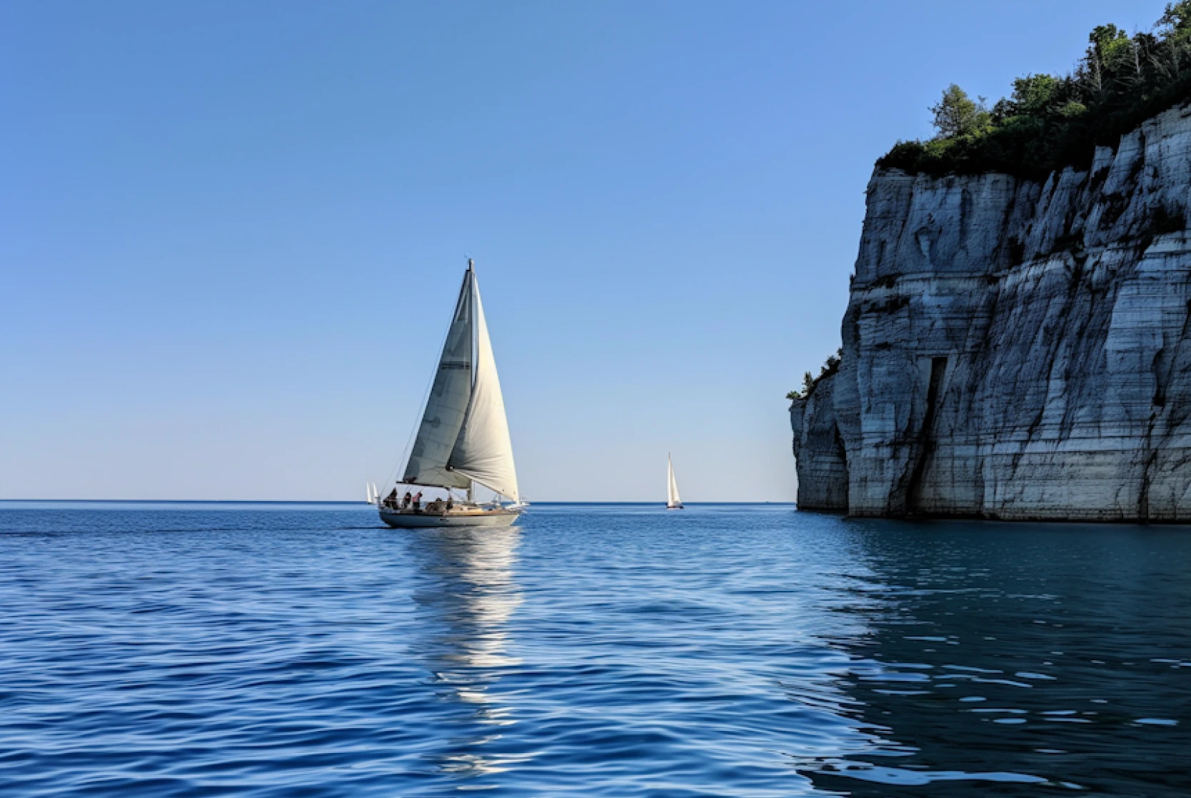 A sailboat with a white sail glides across calm blue water near a tall, rocky cliff. In the background, another smaller sailboat sails by under clear blue skies.