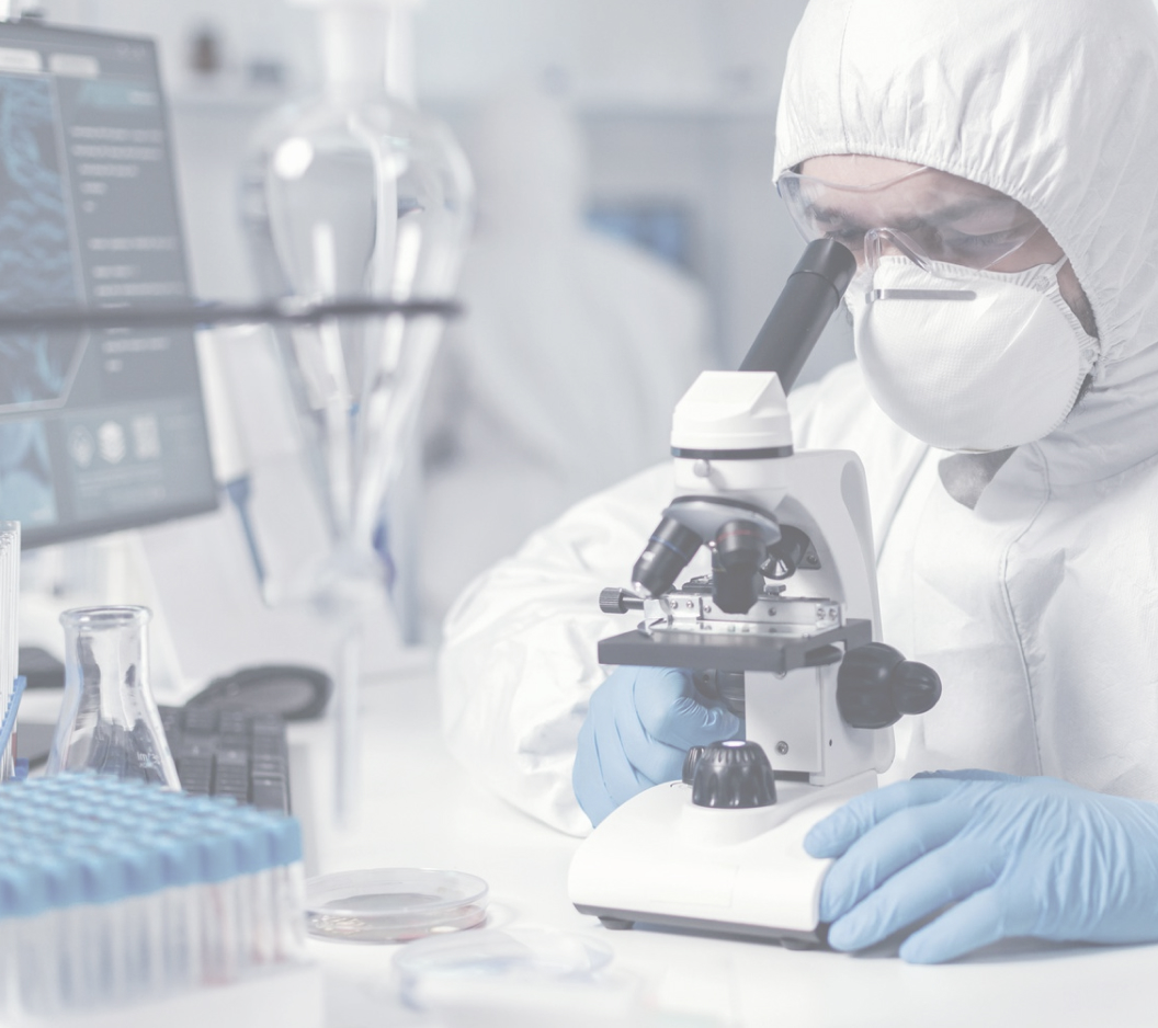 A scientist in a white protective suit and gloves examines a sample through a microscope in a laboratory. Surrounding them are various lab equipment, including test tubes and a computer displaying scientific data.