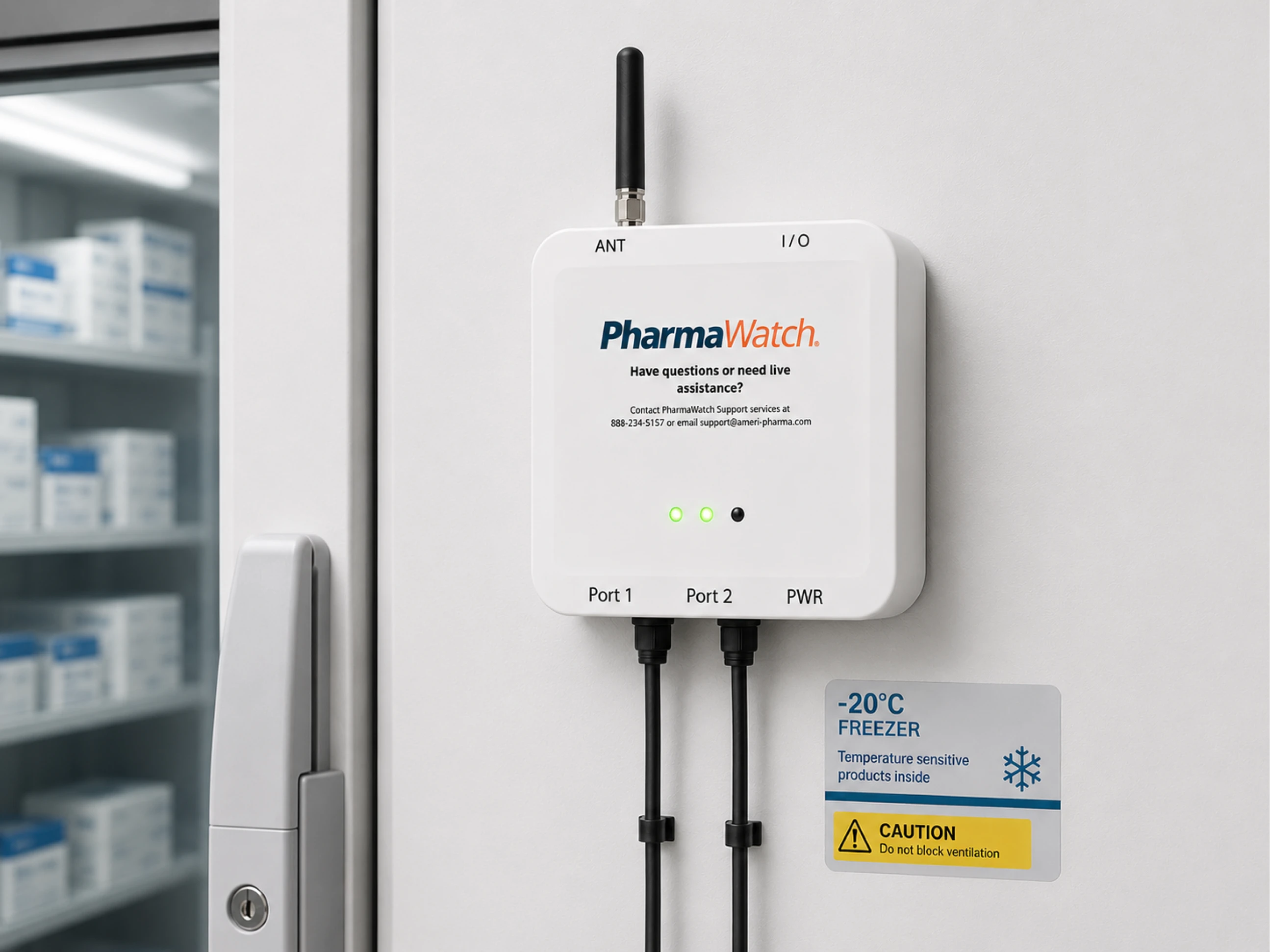 A white PharmaWatch monitoring device is mounted on a wall next to a large gray freezer. The device features an antenna, three green indicator lights, and two ports with black cables connected. A caution sign next to it indicates the freezer temperature is set to -20°C.