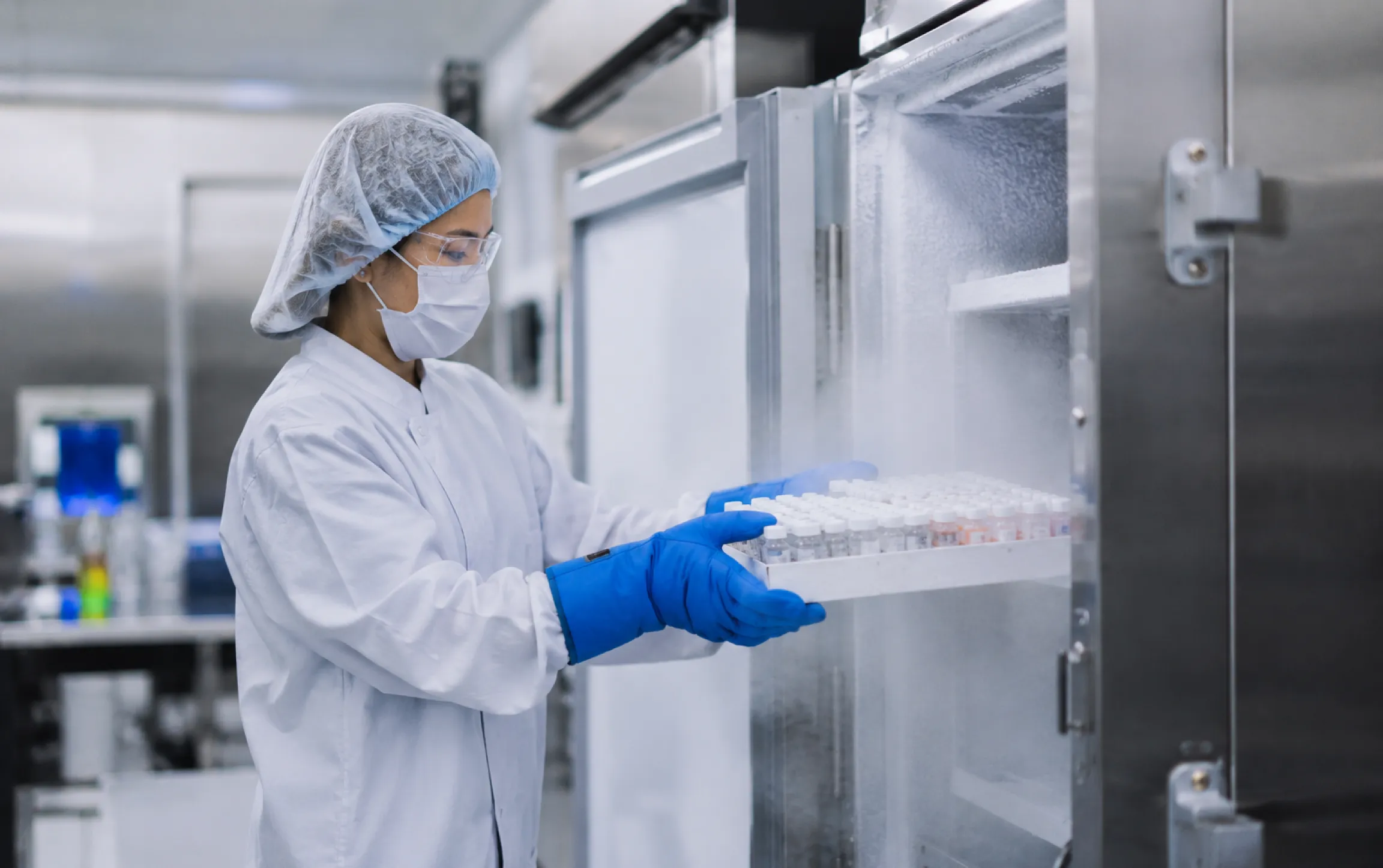 A laboratory technician in a white lab coat, blue gloves, and a hairnet is placing a tray of vials into a large cold storage freezer. The environment appears sterile with stainless steel surfaces and scientific equipment in the background.