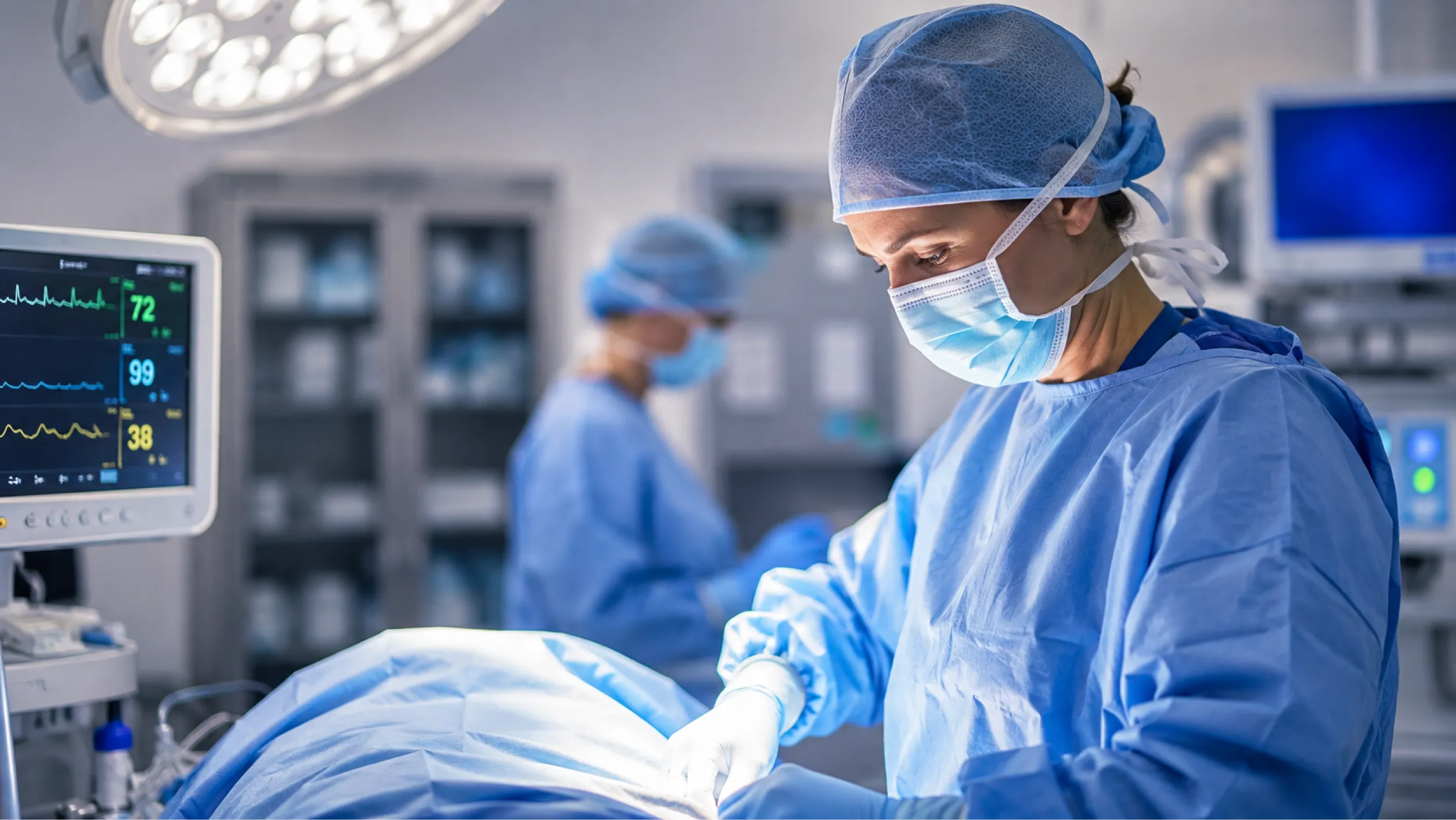 A female surgeon in surgical scrubs and a mask focuses on a surgical procedure with gloves on. In the background, another medical professional is visible, and medical monitors display vital signs.