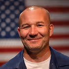 Smiling person in a blue jacket stands in front of a blurred U.S. flag.