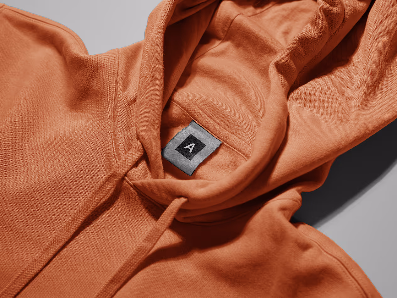 Close-up of an orange hoodie with drawstrings, showing a black inner label with the letter “A.”