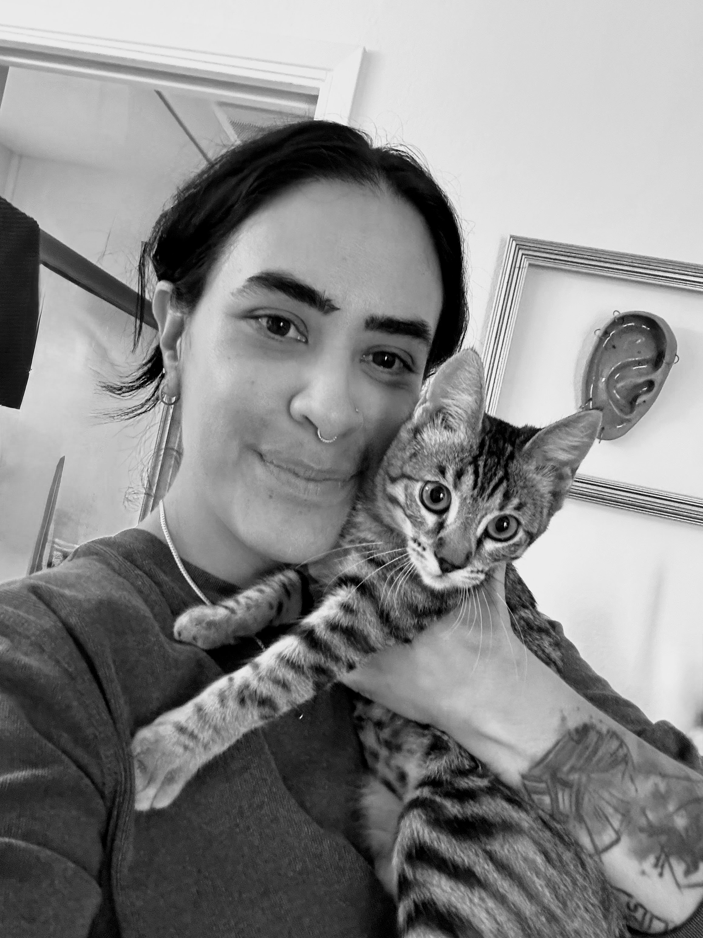 Person with dark hair and nose piercing holding a tabby cat, both looking toward the camera in a home setting.