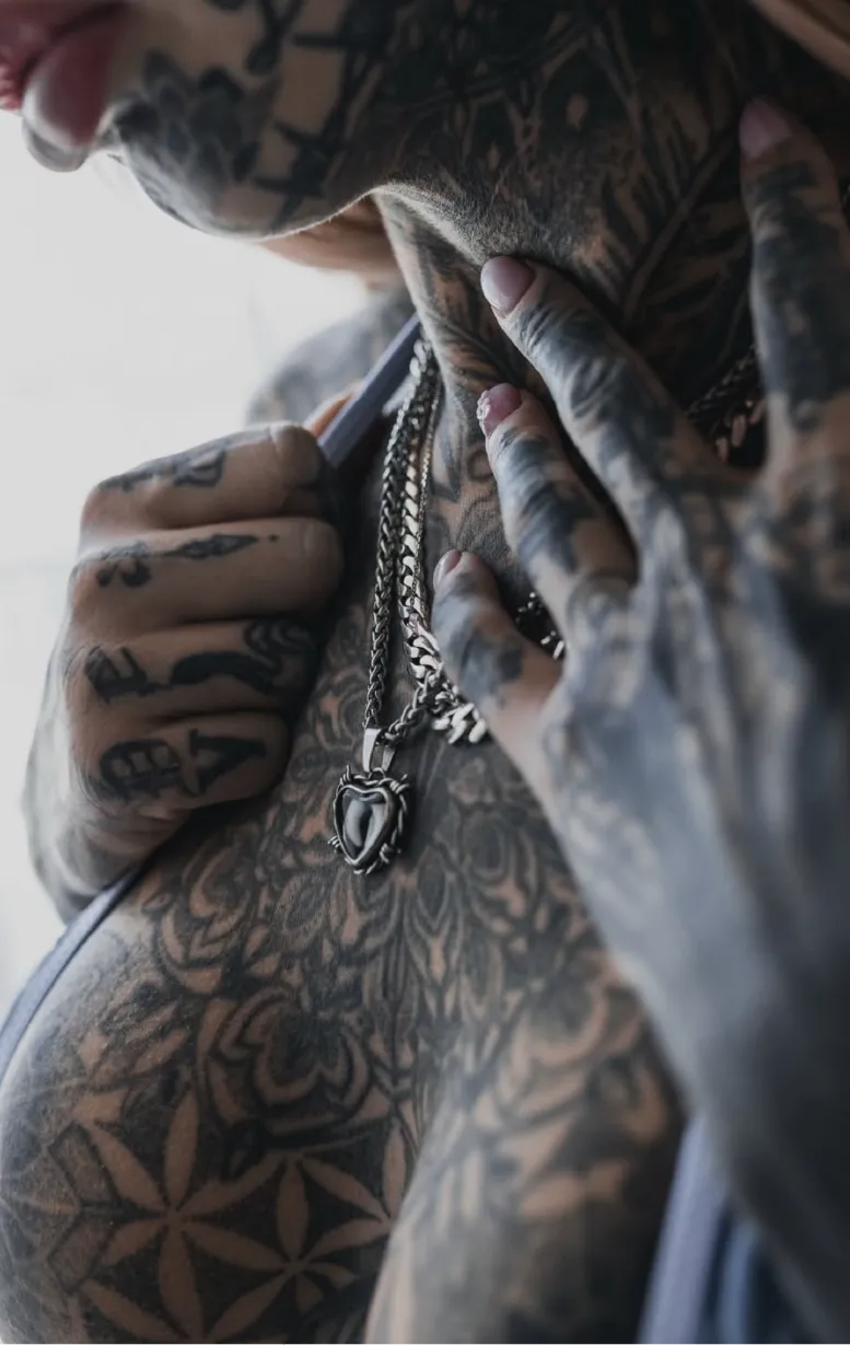 Close-up of tattooed chest and hands with a silver heart-shaped pendant necklace.