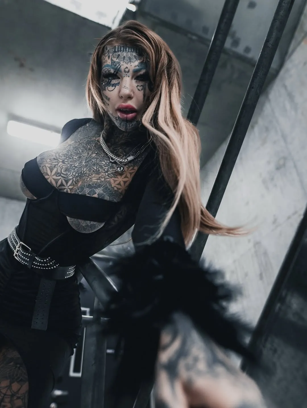 Tattooed woman with long hair wearing a black fitted dress and chains, reaching out towards the camera in a dimly lit industrial setting.