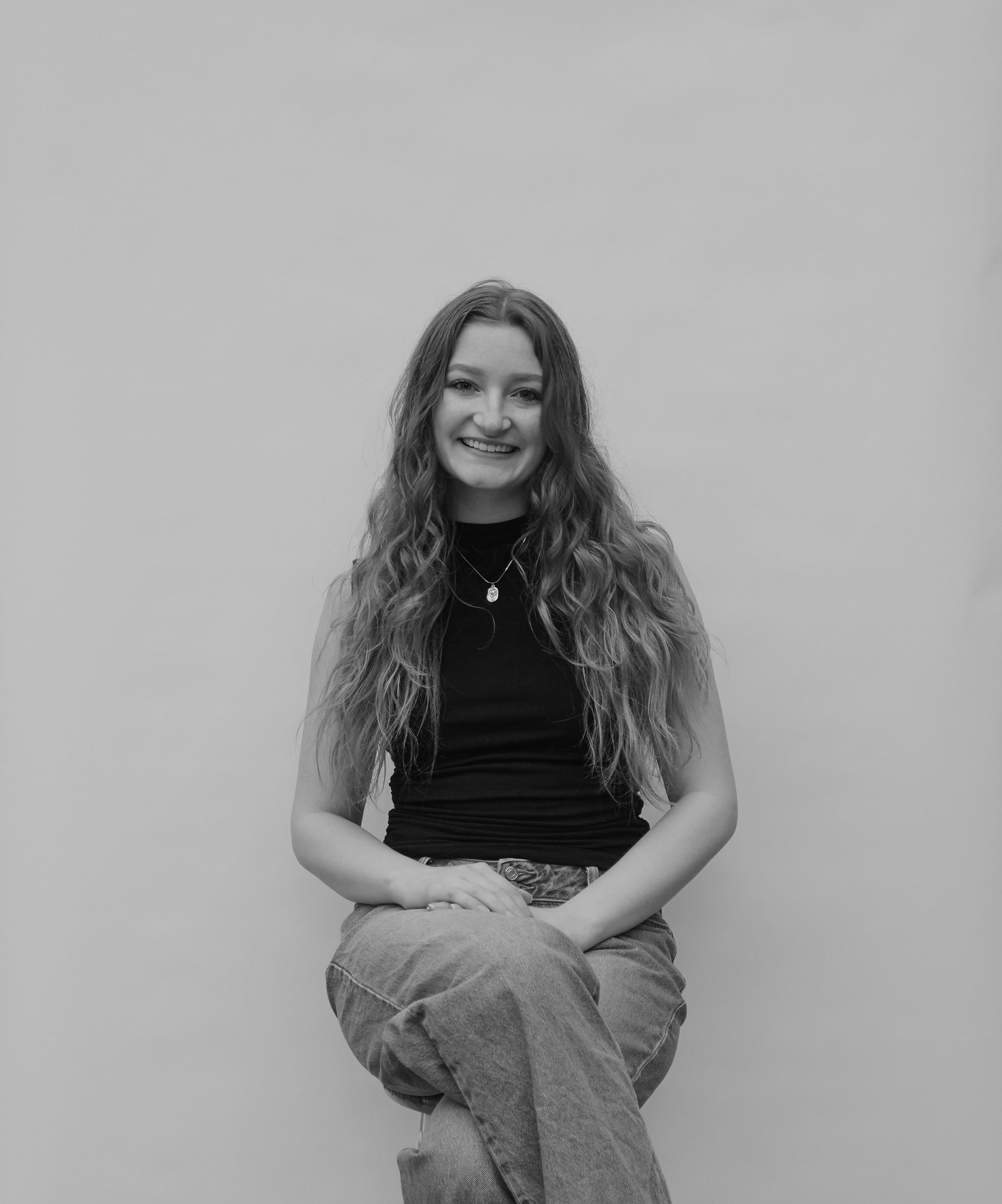Young woman, Ella Janssen, with long wavy hair smiling, seated against a plain background wearing a sleeveless black top and jeans.