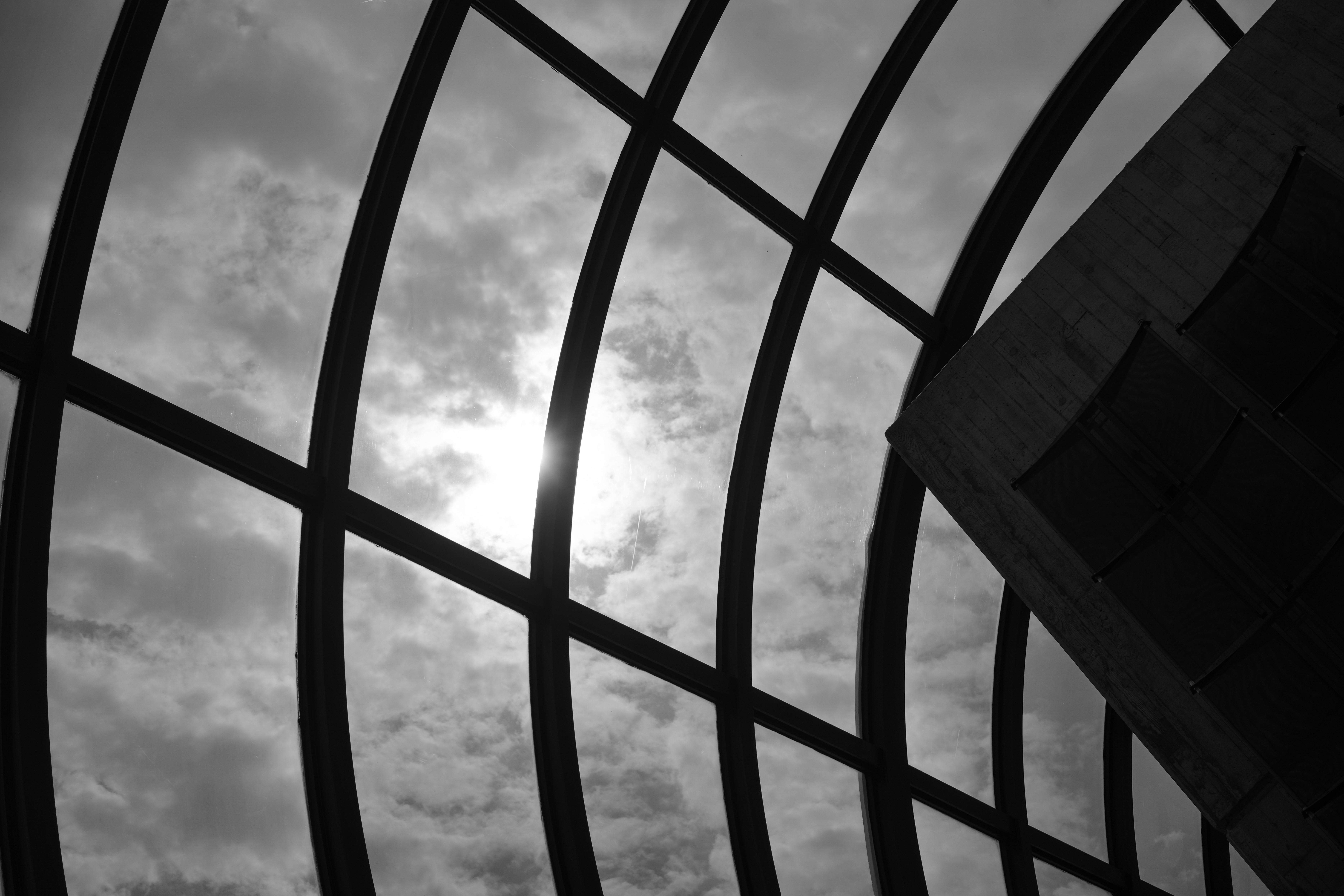 Black and white photo of curved window frames with the sun and cloudy sky visible through the glass.