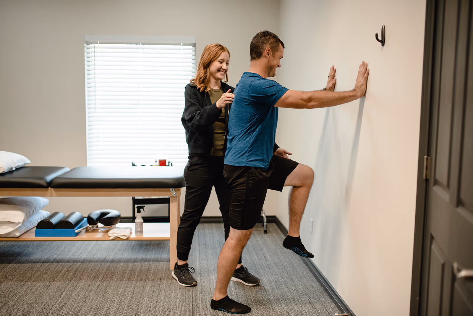 Man in blue shirt doing a wall-supported leg stretch while a woman in black athletic wear assists him in a clinical setting.