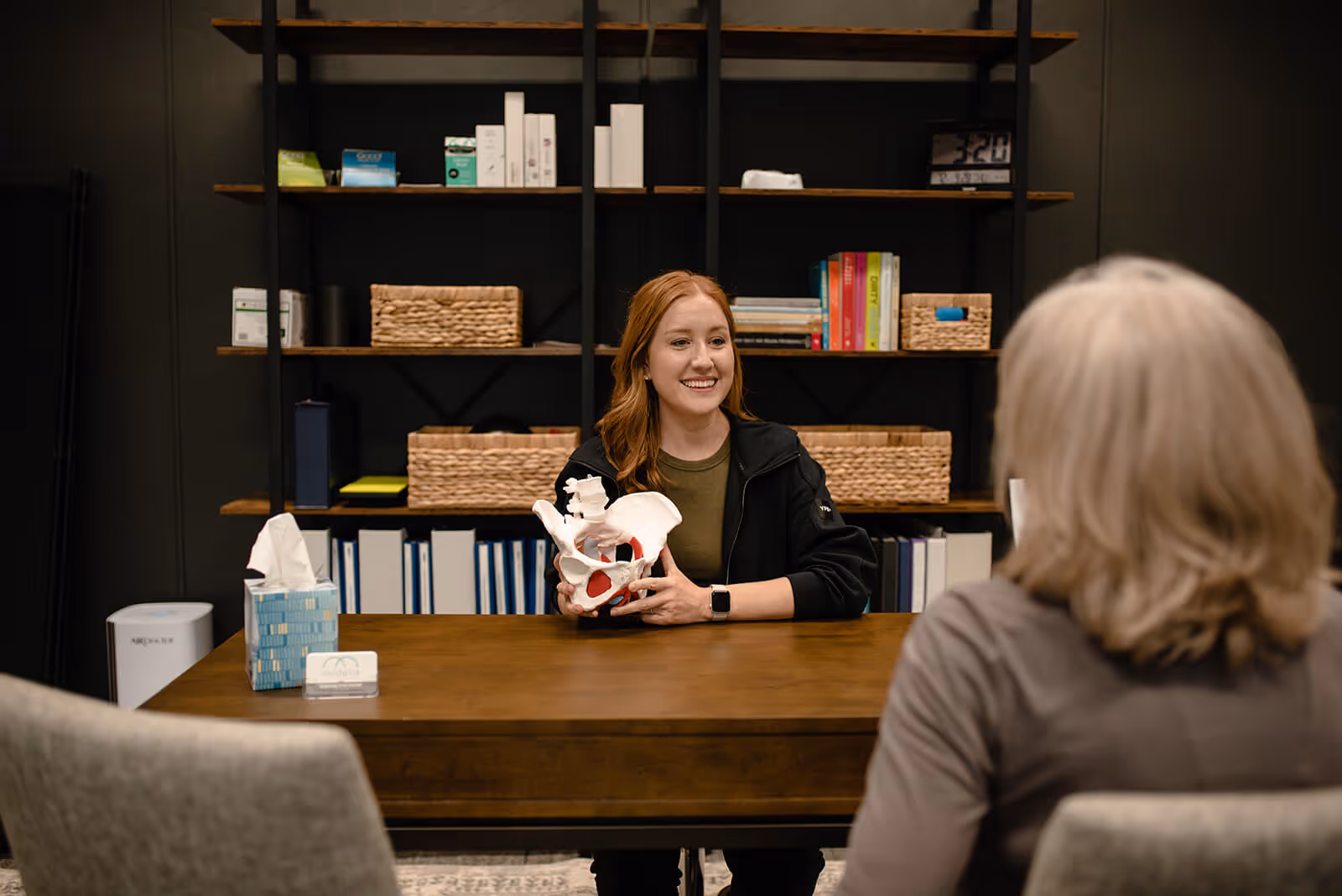 Smiling woman holding a pelvic bone model while sitting at a wooden table, talking to another person in an office with shelves and books in the background.