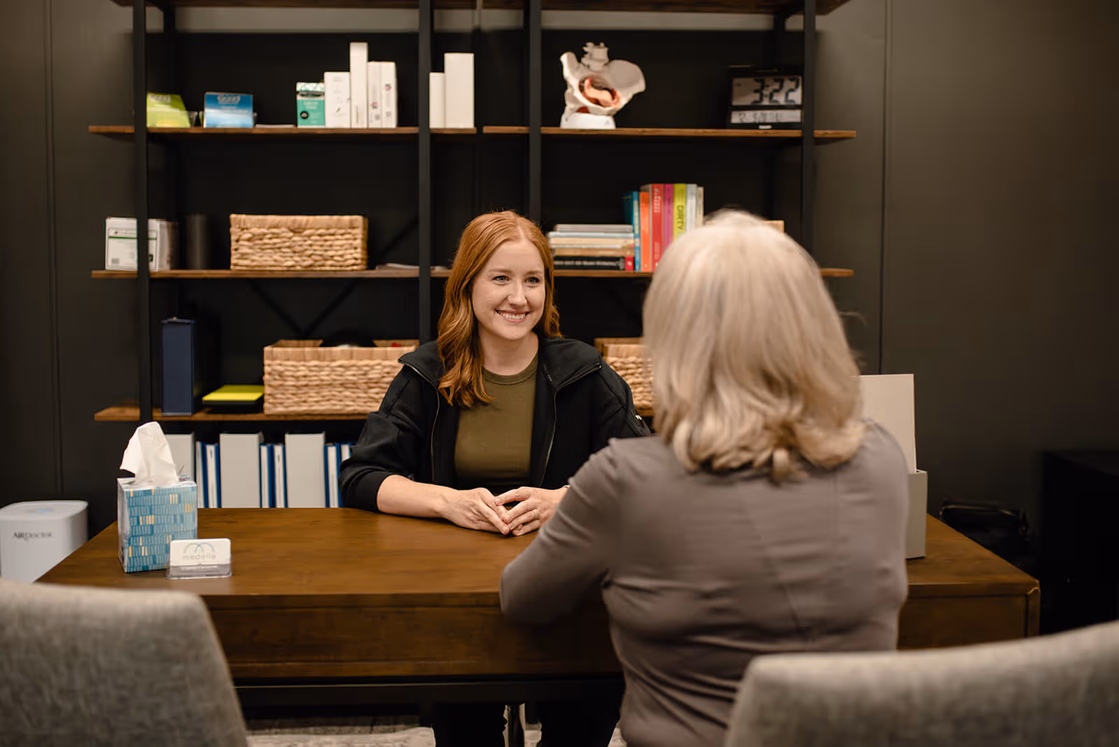 Smiling young woman with red hair sitting at a desk facing an older woman with gray hair in an office setting.