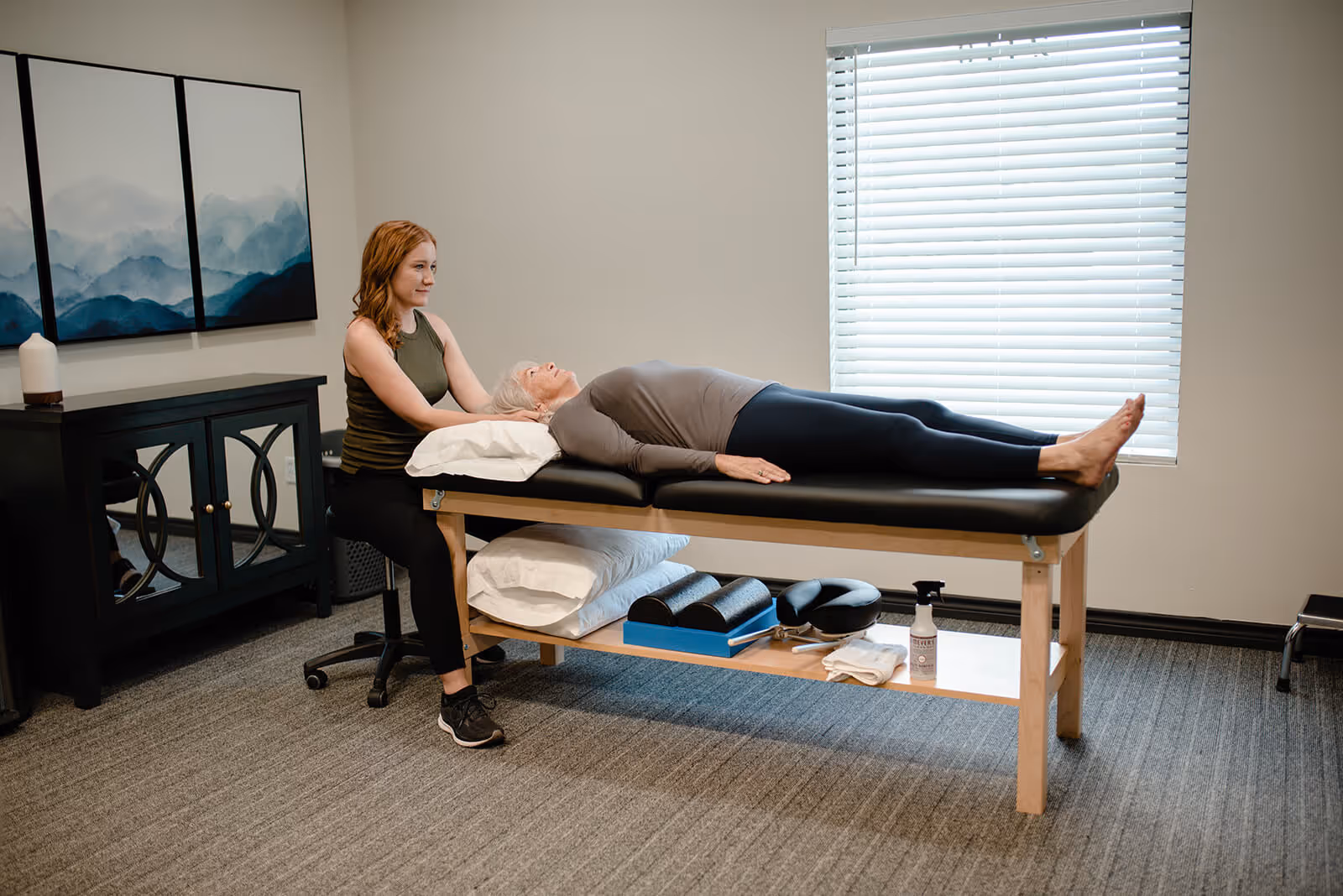 Therapist kneeling beside a treatment table, gently supporting the head of a woman lying down in a therapy room.