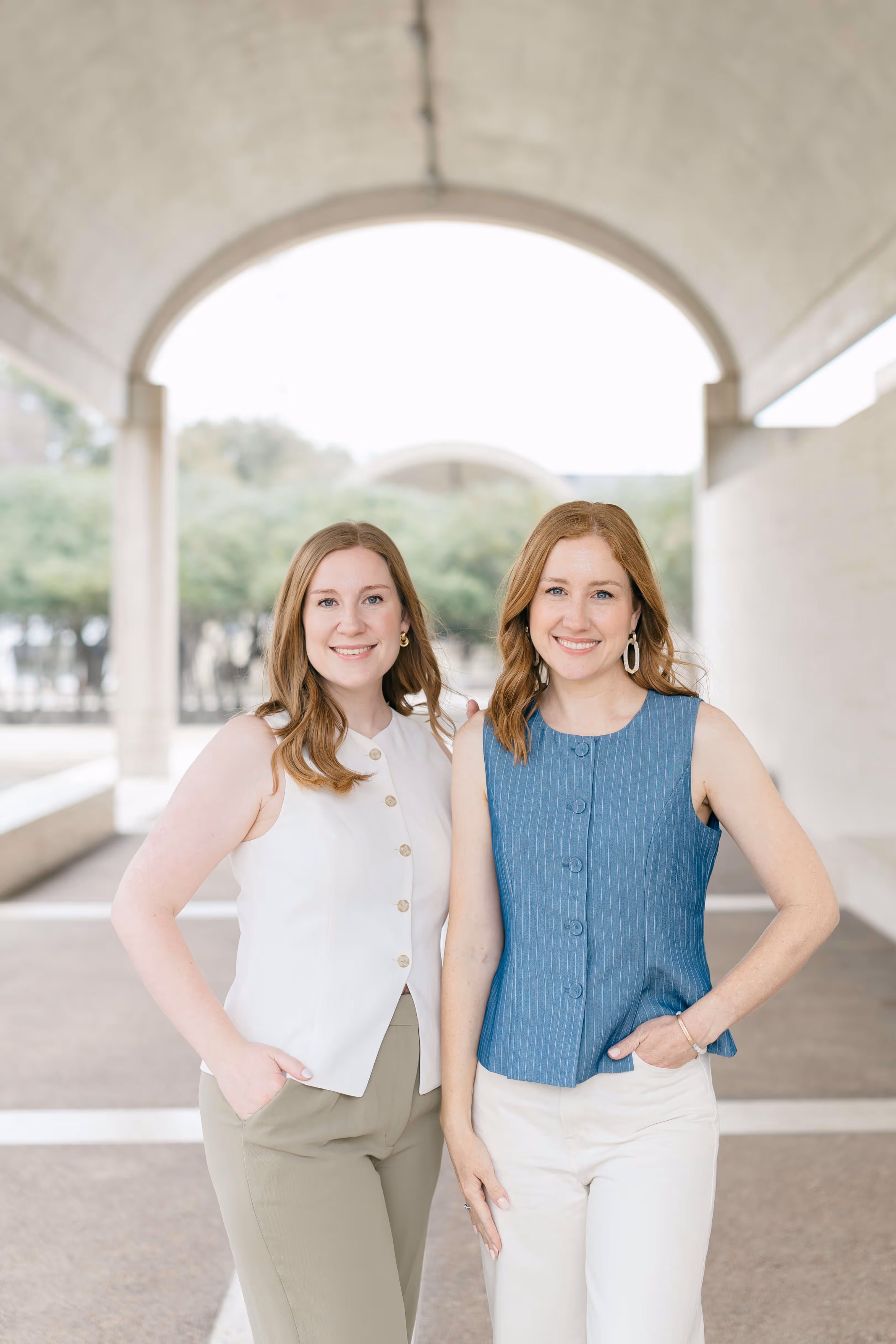 Two smiling women standing side by side under an archway, one wearing a sleeveless white top and beige pants, the other in a sleeveless blue top and white pants.