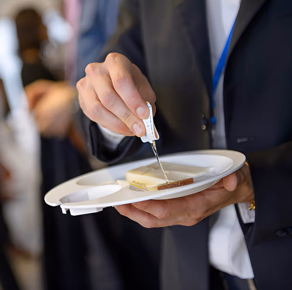 Person in a suit holding a plate and serving a small portion of cheese with a cheese slicer.
