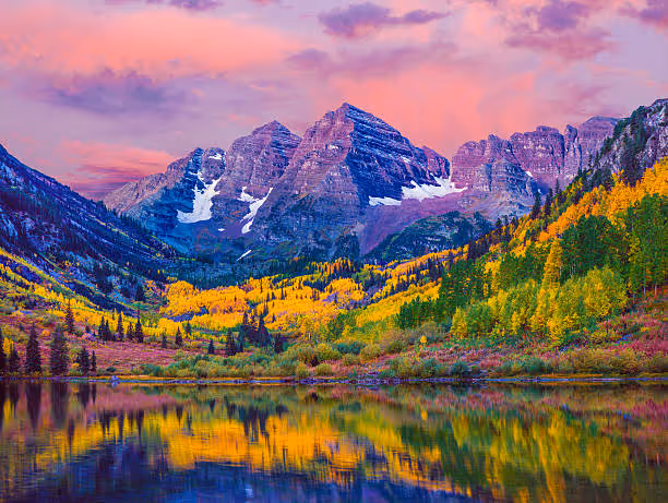 Reflection of fall-colored trees and rugged snow-capped mountains at sunset over a calm lake.