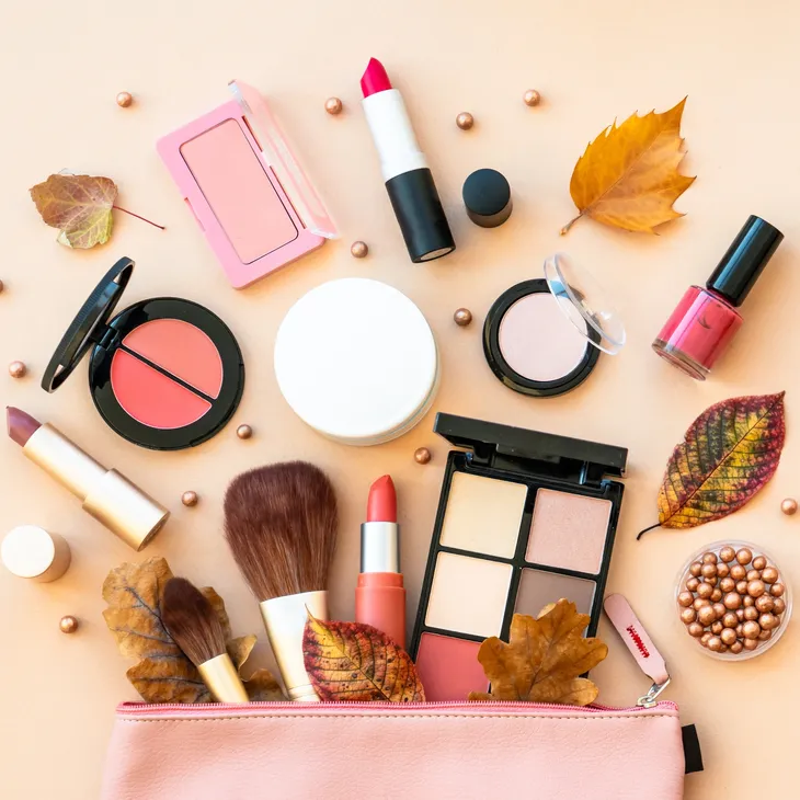Assorted cosmetics including lipsticks, brushes, blush, eyeshadow, and nail polish with autumn leaves and pearls.