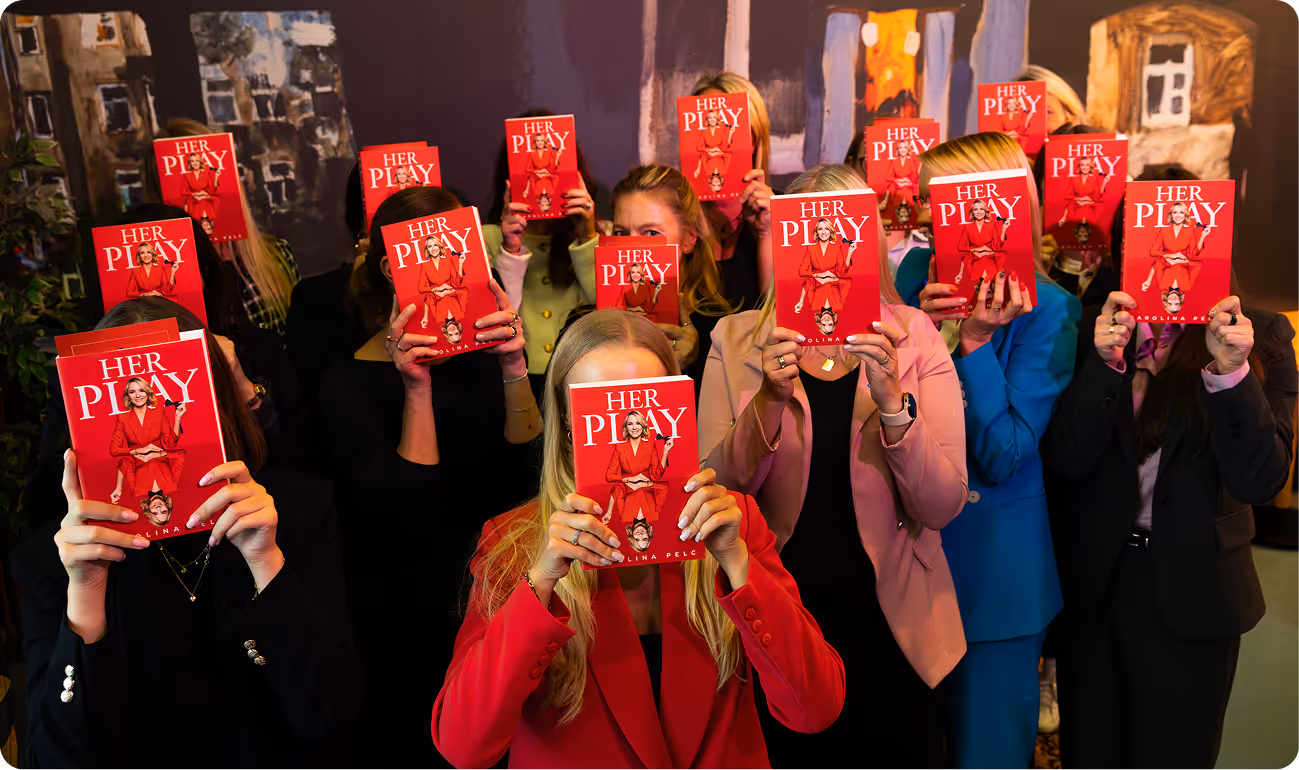 Group of women holding up red books titled 'HER PLAY' with a woman in a red suit on the cover, covering their faces.