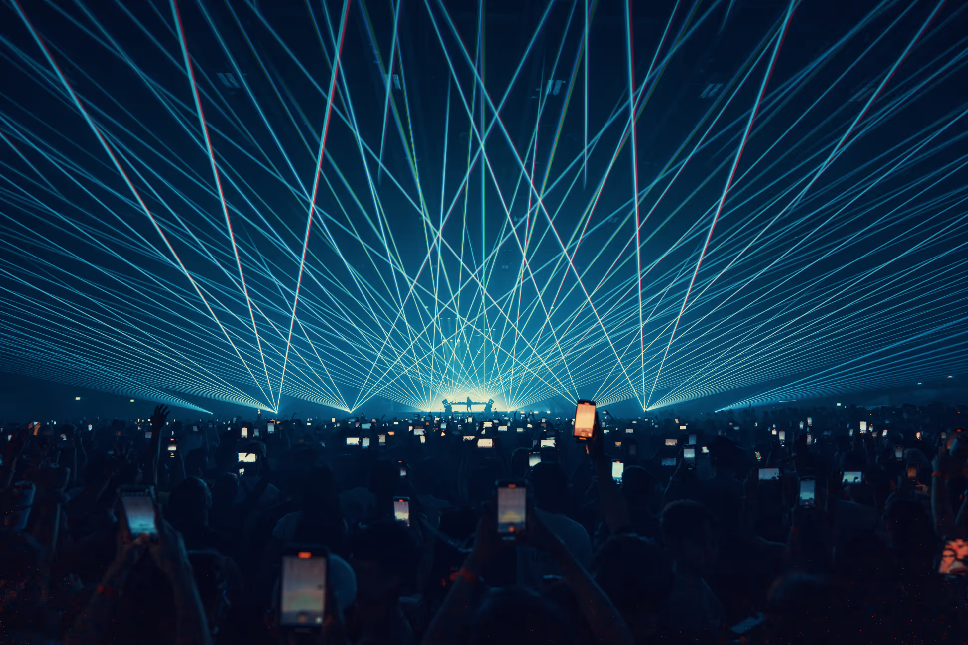 Large crowd at a concert with many holding up phones capturing a light show with blue and green laser beams.