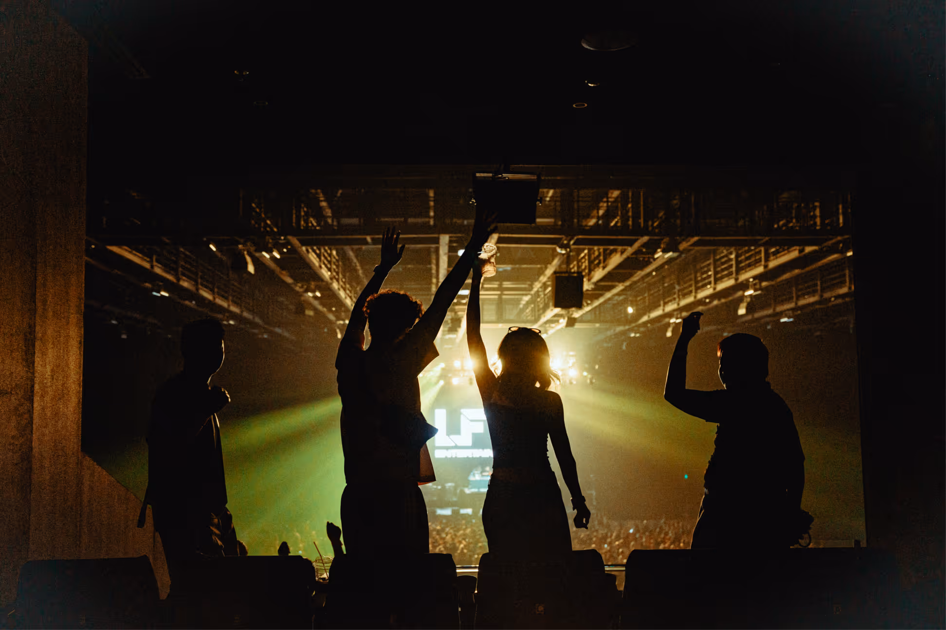 Silhouettes of four people raising their arms and celebrating at an indoor concert with bright stage lights in the background.