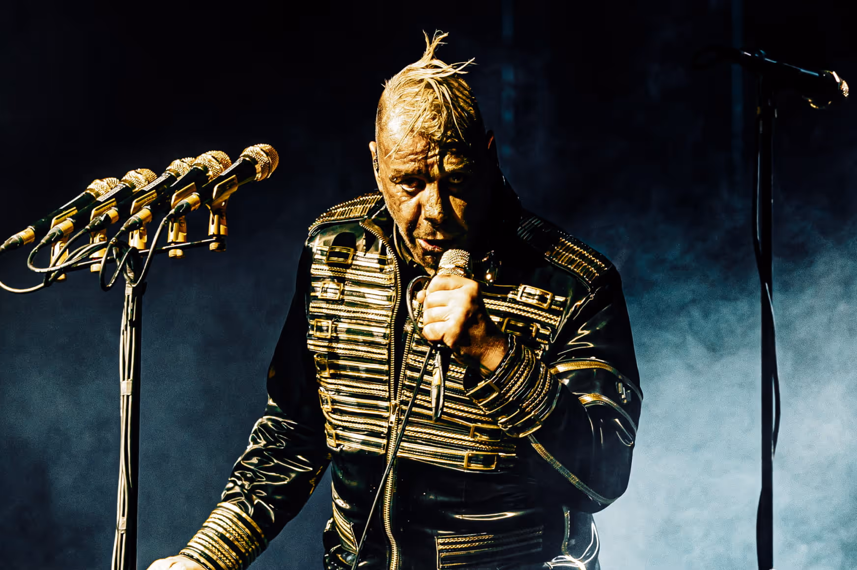 Male singer with blonde mohawk and detailed black and gold jacket performing with multiple golden microphones on stage.
