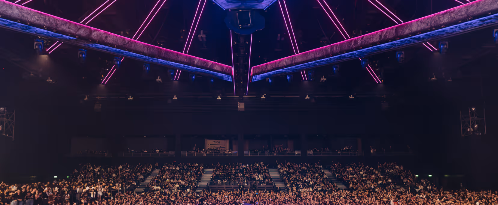 Large crowd gathered inside a dimly lit arena with pink neon lights on the ceiling.