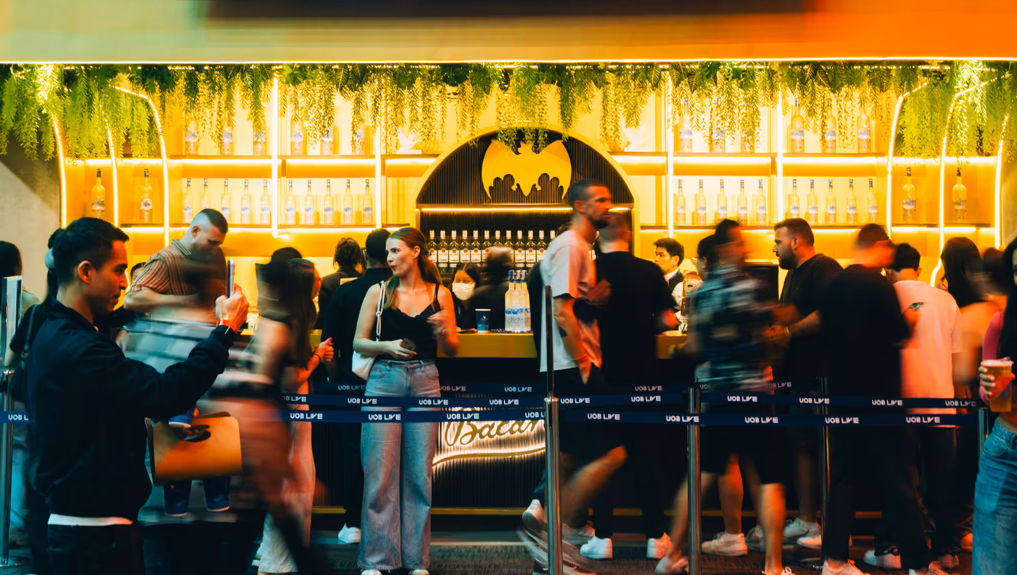 Crowd of people standing and moving inside a brightly lit bar with illuminated shelves of bottles and hanging greenery.