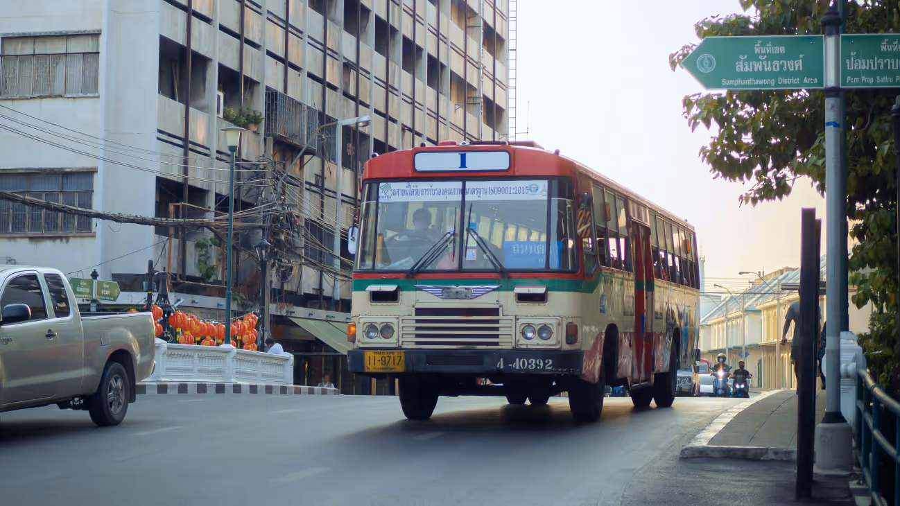 Red and green public bus driving on a city street with a silver pickup truck on the left and motorcyclists in the background.