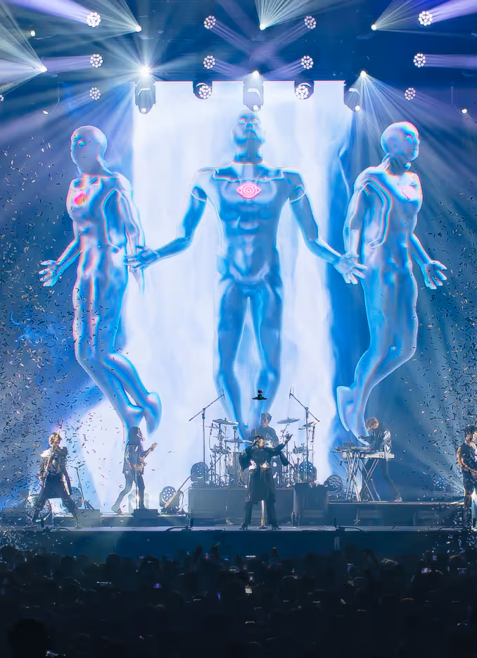 Concert stage with a band SLOT MACHINE performing in front of three large glowing humanoid figures projected on a screen, with bright spotlights and confetti.