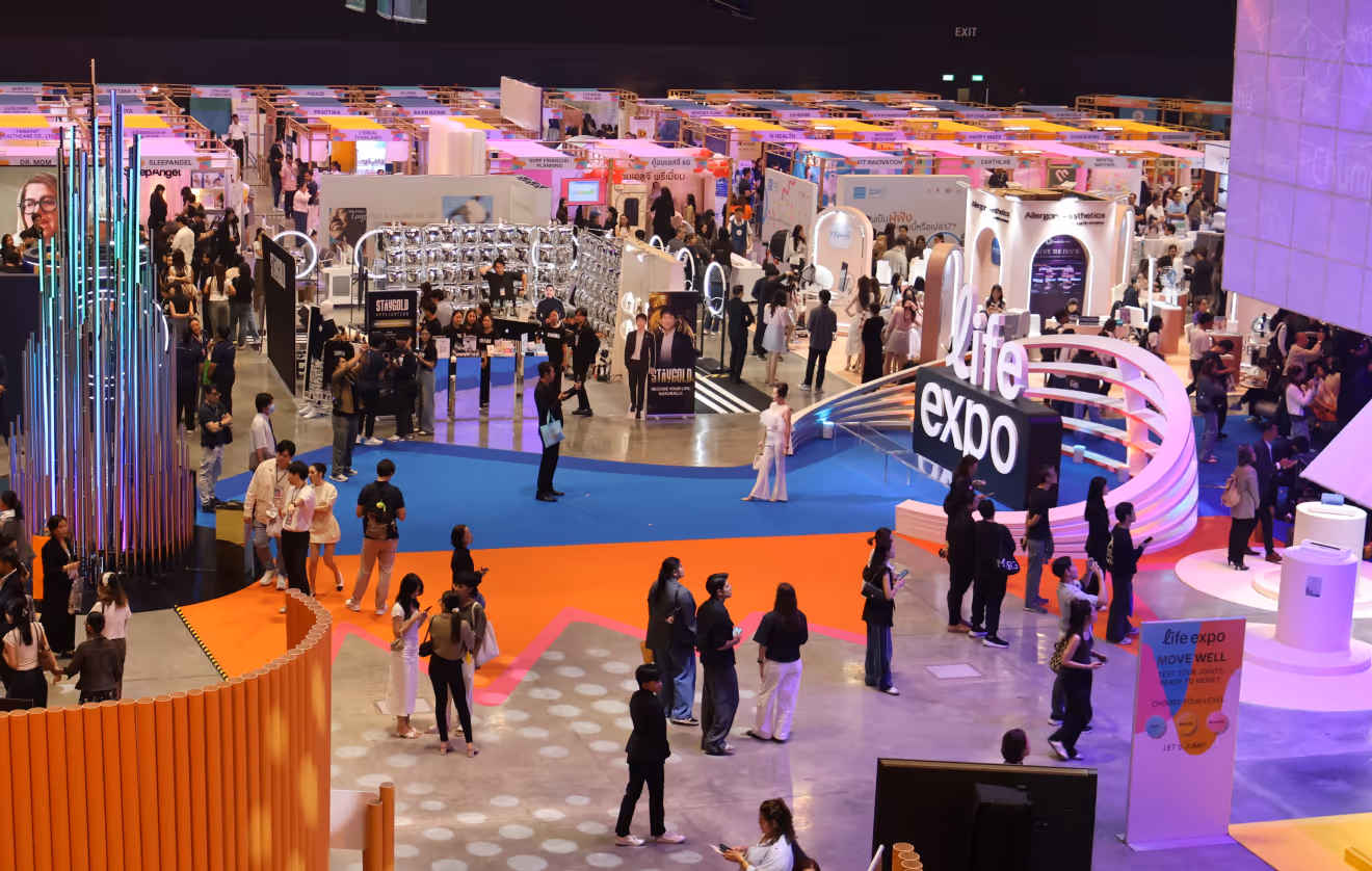 Large indoor life expo event with numerous booths, colorful flooring, and many attendees walking and interacting.