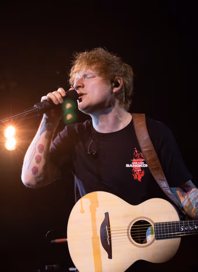 Singer Ed Sheeran passionately singing into a microphone on stage, holding an acoustic guitar at UOB LIVE Grand Opening.