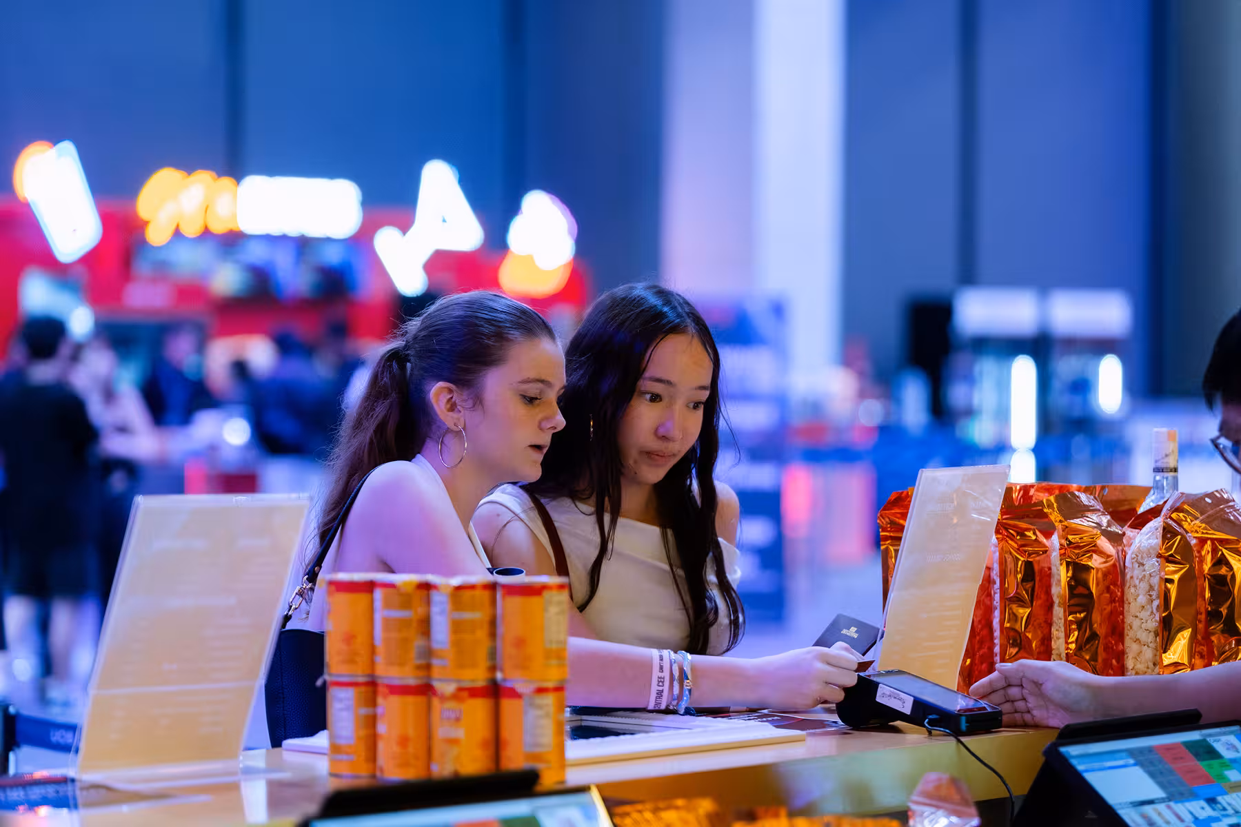 Two women using a credit card machine at a counter with cans and bags of popcorn in a brightly lit UOB LIVE Concession at Prefunction Area
