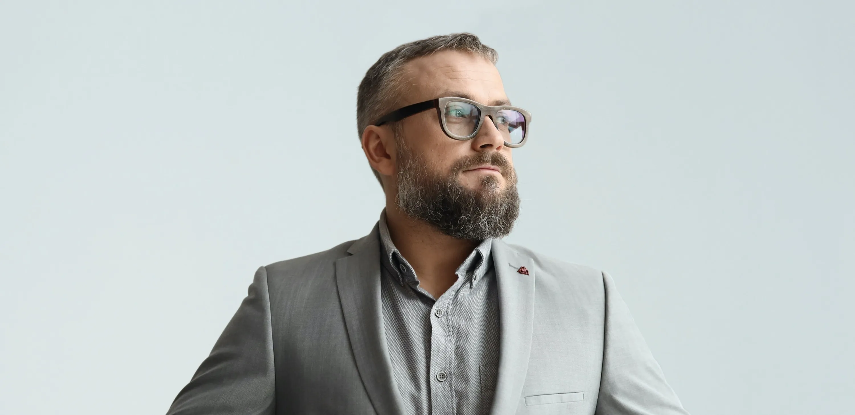 Bearded man wearing glasses and a light gray suit buttoning his jacket while looking to the side.