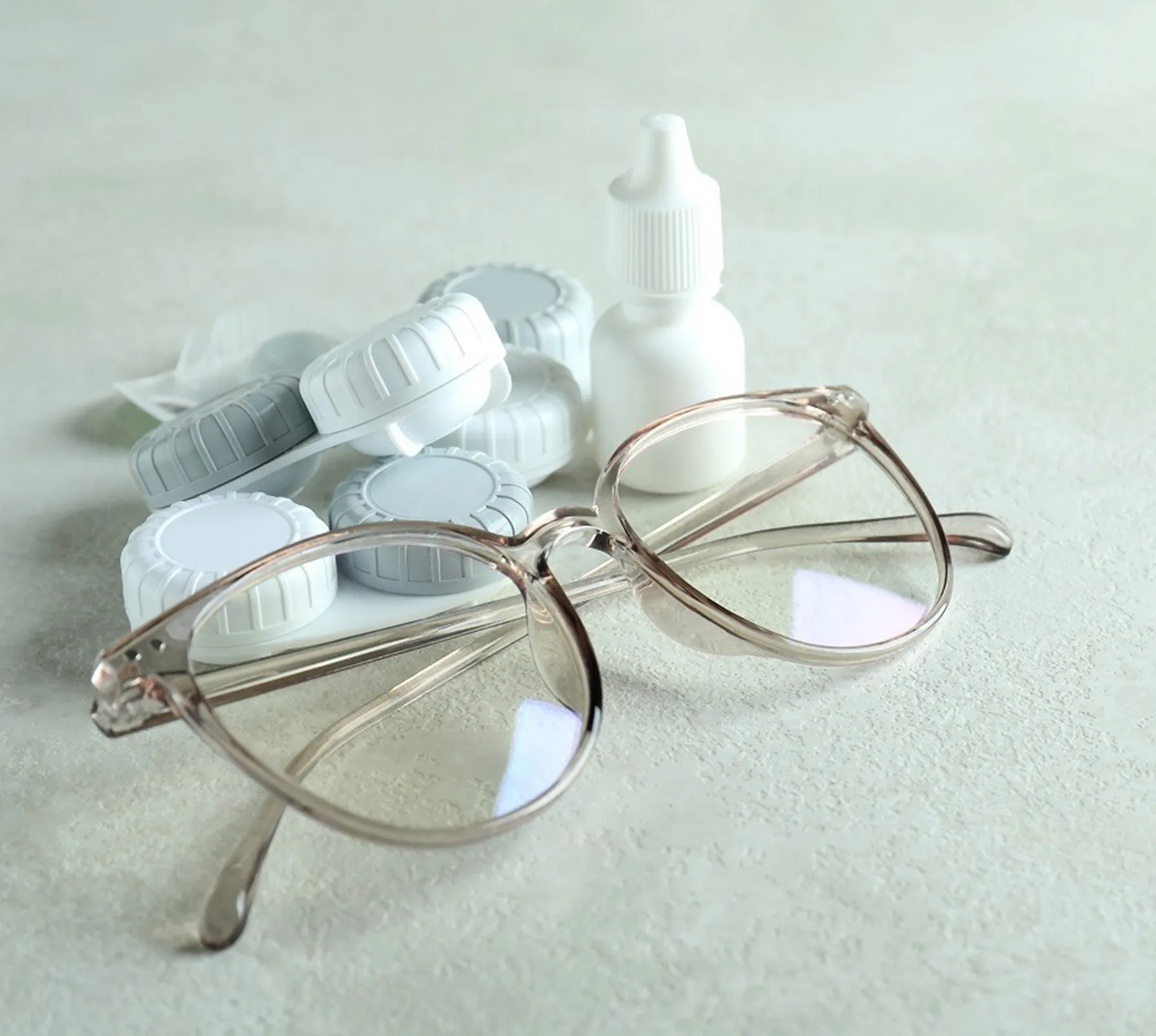 Clear eyeglasses placed near a contact lens case and a small white eye drop bottle on a light surface.