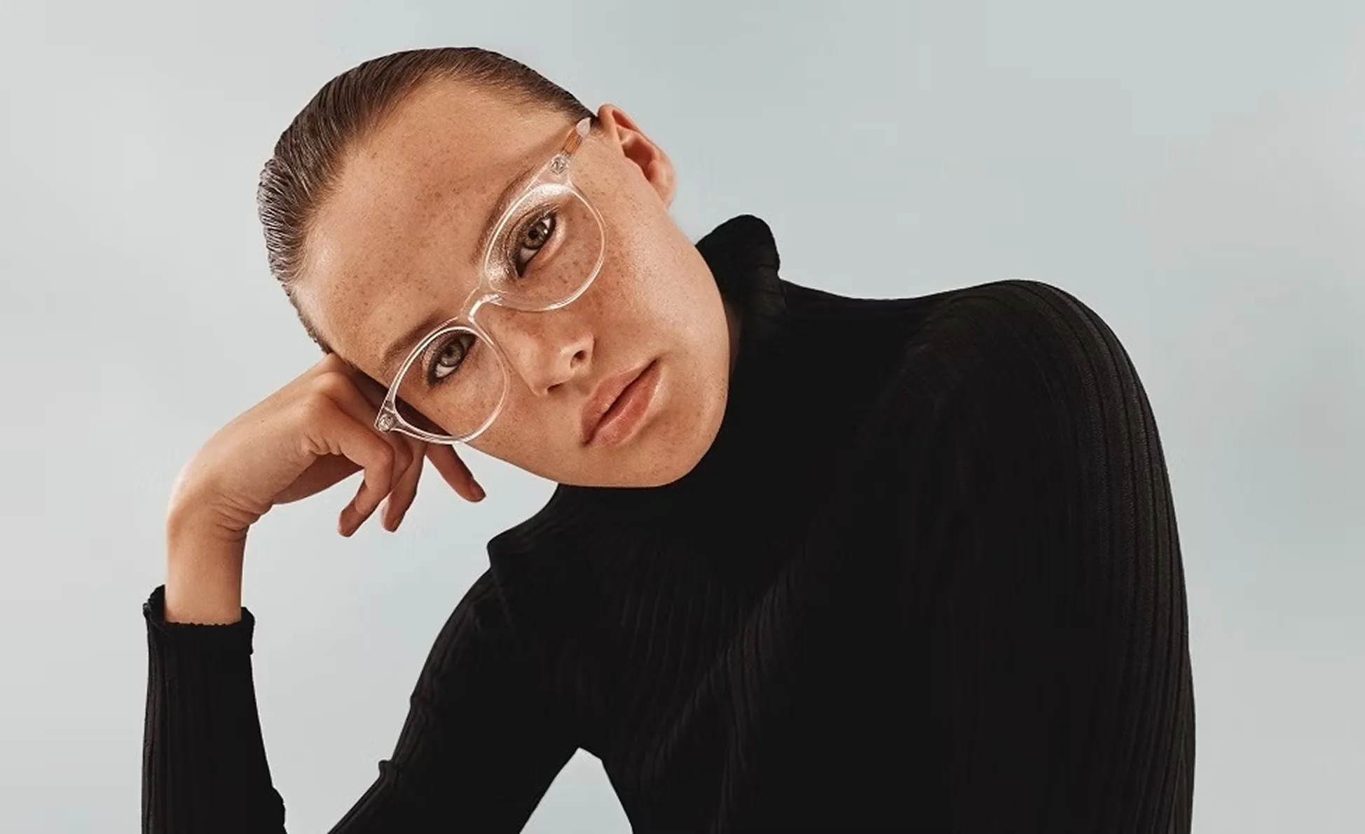 Young woman with slicked-back hair wearing clear glasses and a black turtleneck sweater resting her head on her hand.