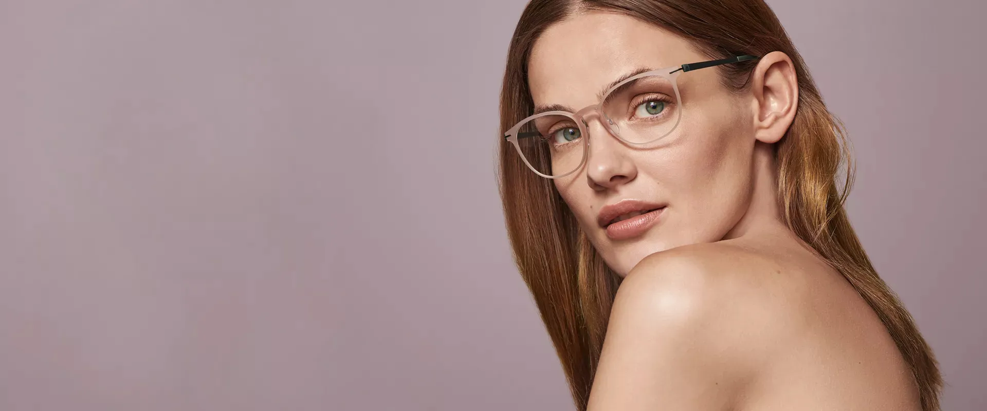 Close-up of a woman with light brown hair wearing translucent eyeglasses against a muted mauve background.