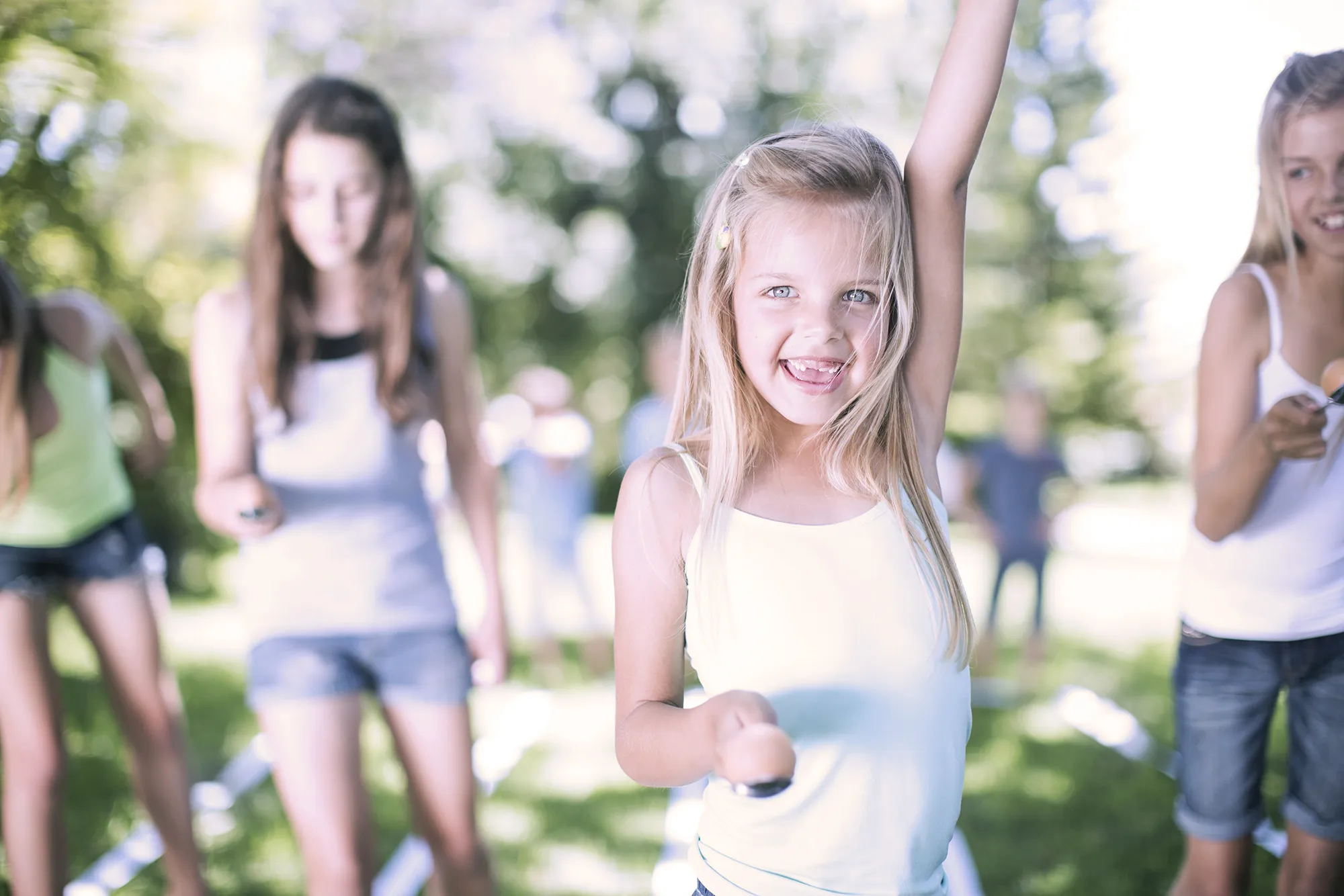 Young girl balancing an egg on a spoon with her arm raised, smiling excitedly outdoors with other children in the background.