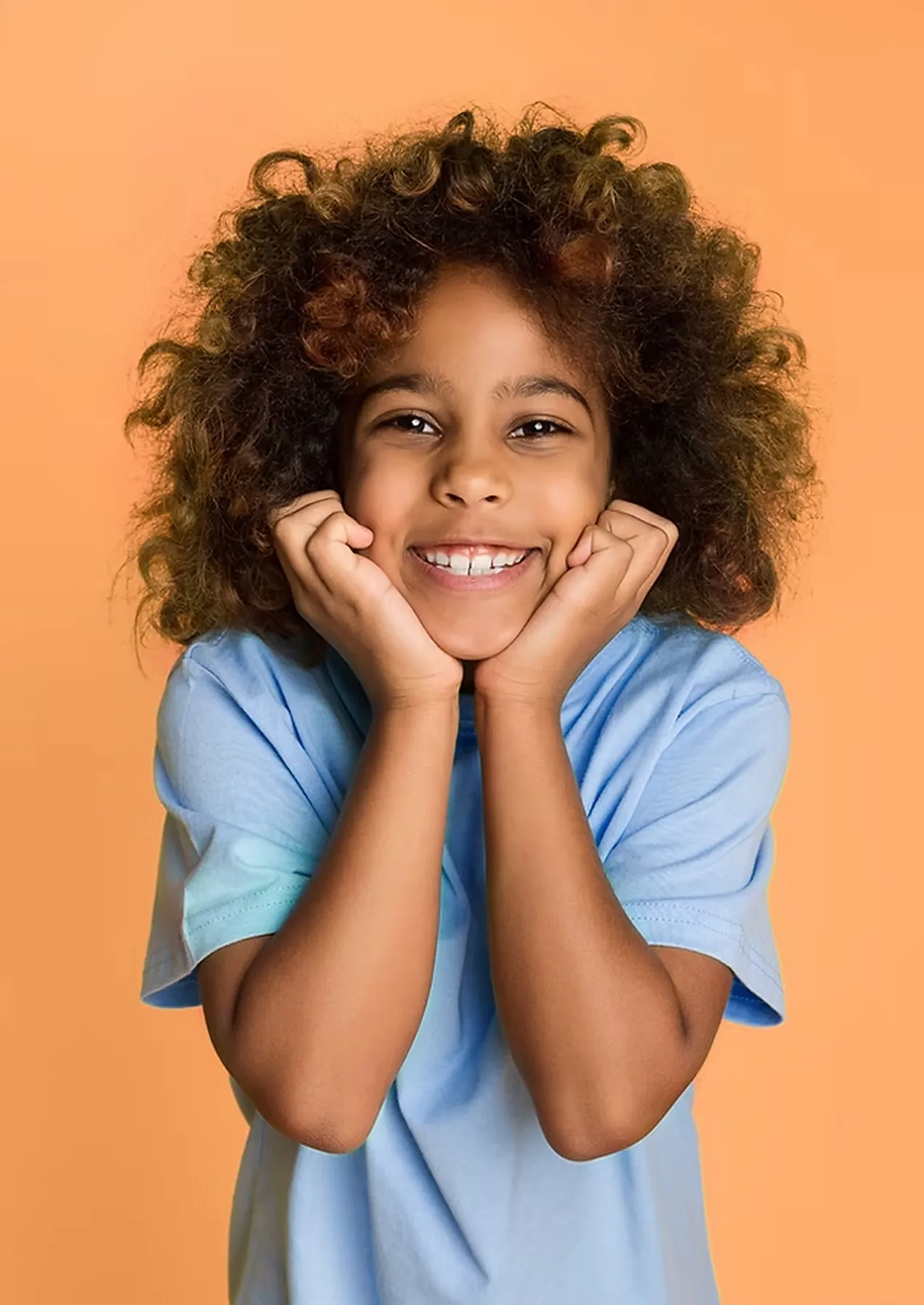 Smiling child with curly hair wearing a light blue shirt, resting face in hands against a peach background.