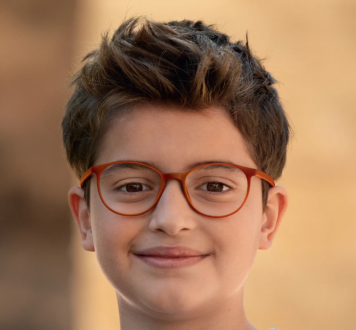 Smiling young boy with short dark hair and orange glasses against a blurred beige background.