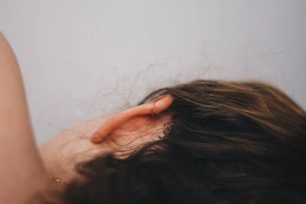 Close-up of a person's ear with a small gold hoop earring and dark brown hair against a light gray background.