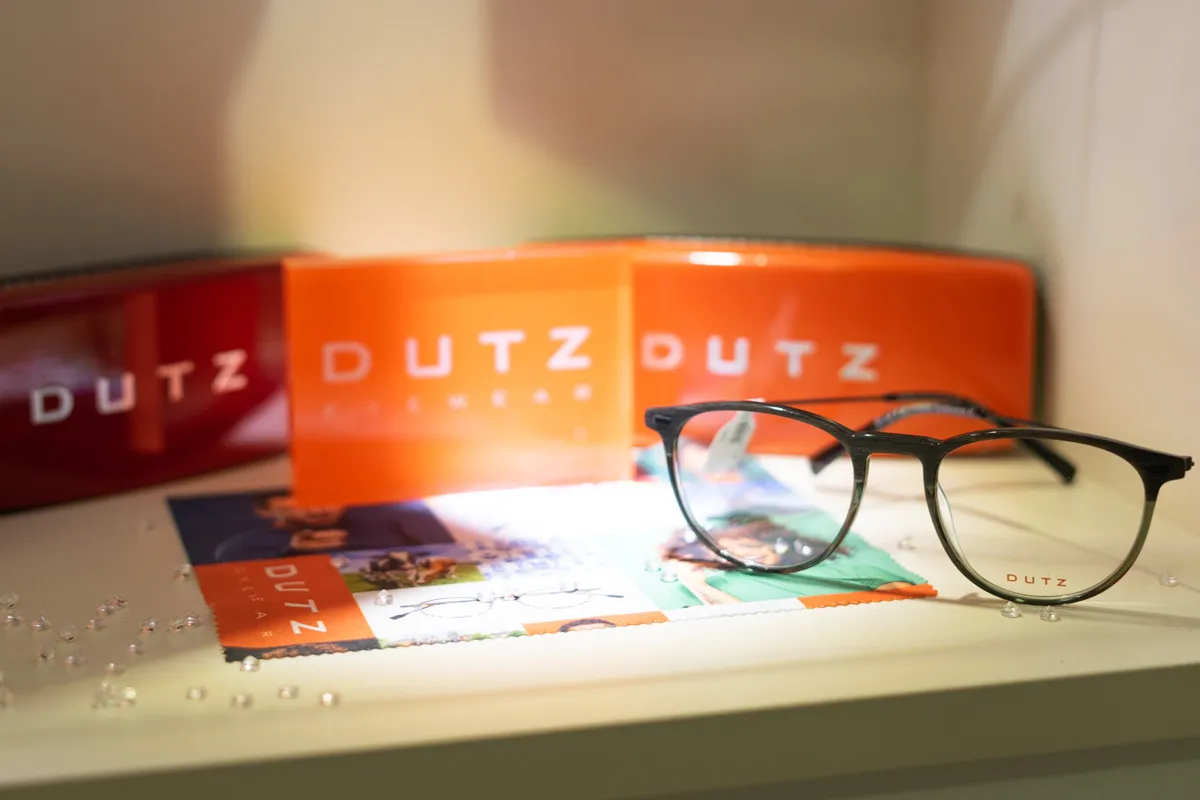 Black-framed DUTZ eyeglasses on a surface with DUTZ packaging and promotional materials in the background.