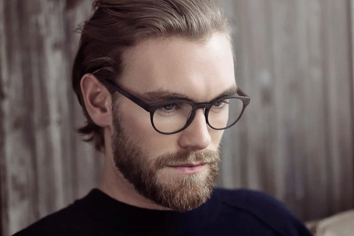 Young man with beard and glasses looking downward, wearing a dark sweater against a blurred wooden background.