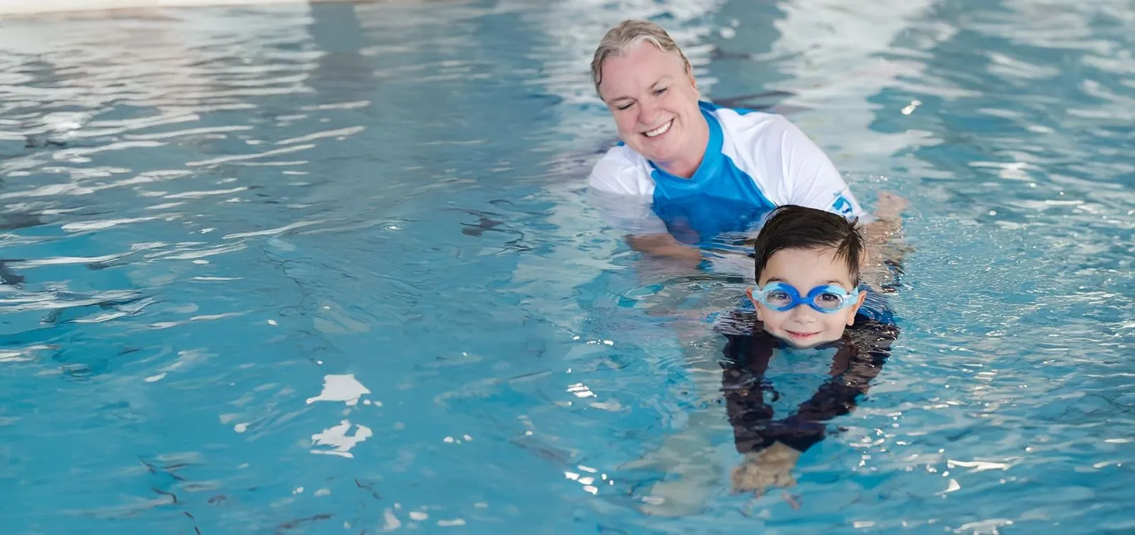 A boy in goggles holding a kickboard at the edge of the pool during a swimming lesson