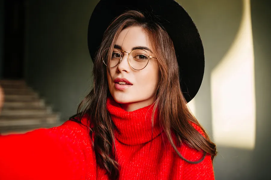 Young woman with long brown hair wearing glasses, a black hat, and a bright red turtleneck sweater taking a selfie indoors.