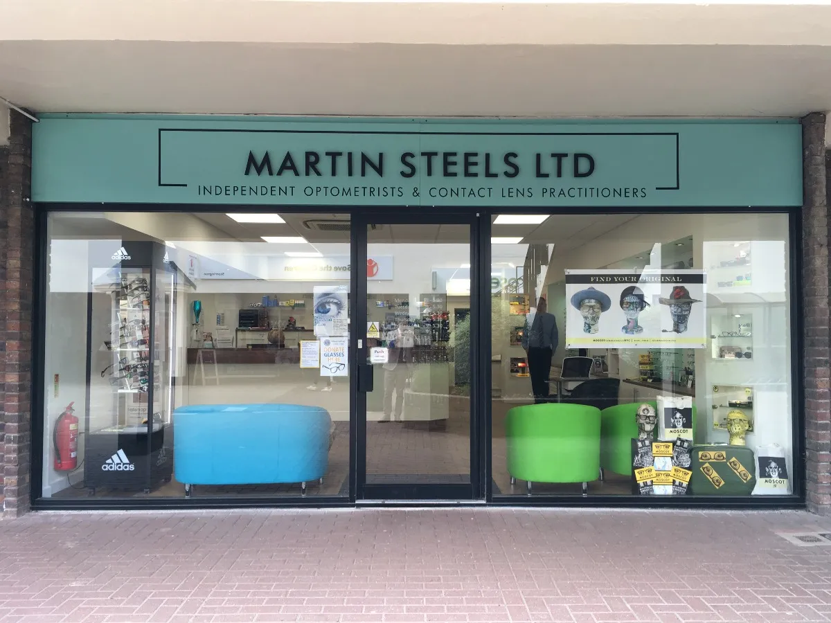 Storefront of Martin Steels Ltd optometrists with blue and green chairs, eyewear display, and promotional posters inside.