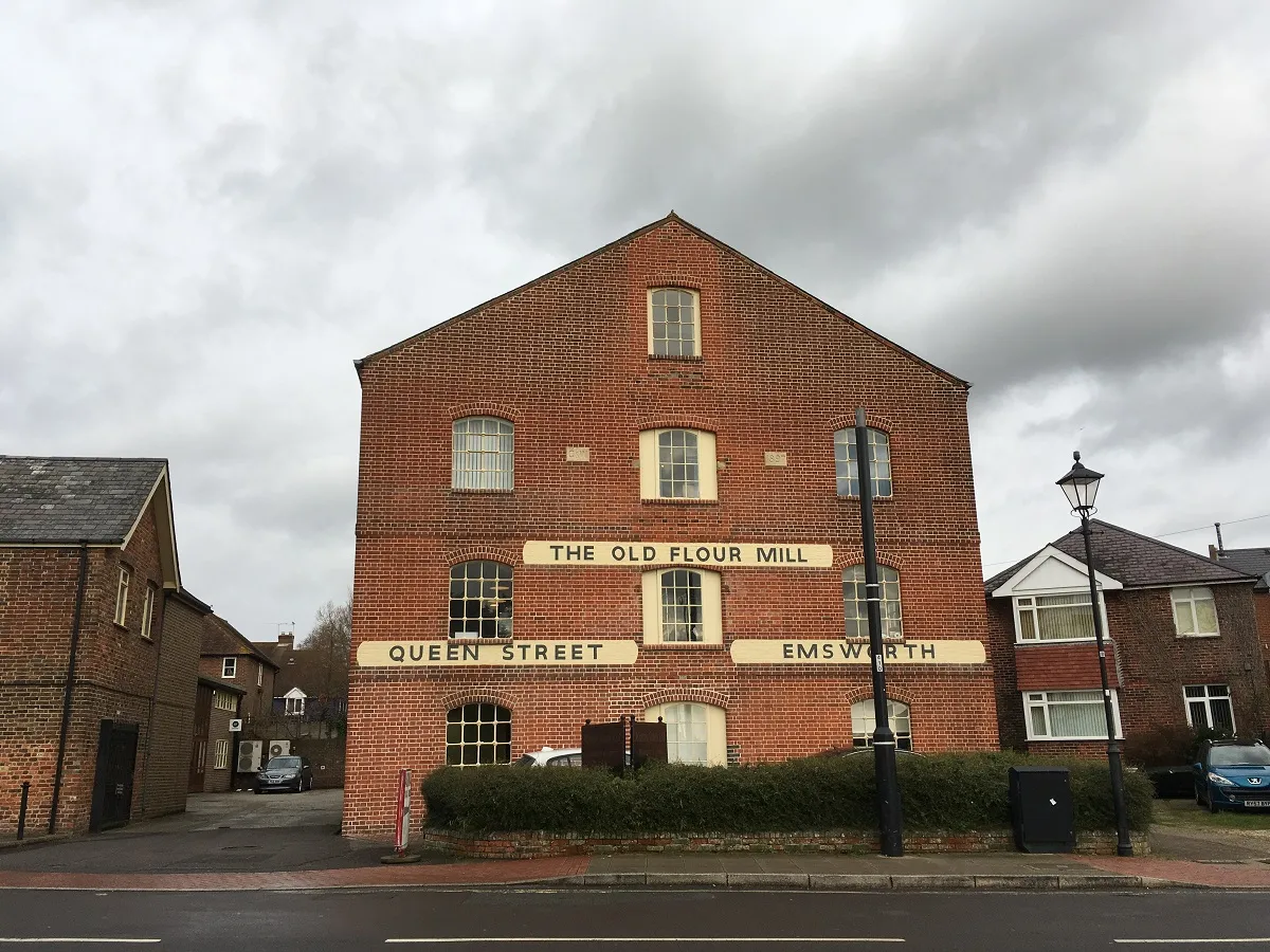 Red brick building with signs reading 'The Old Flour Mill,' 'Queen Street,' and 'Emsworth' under a cloudy sky.