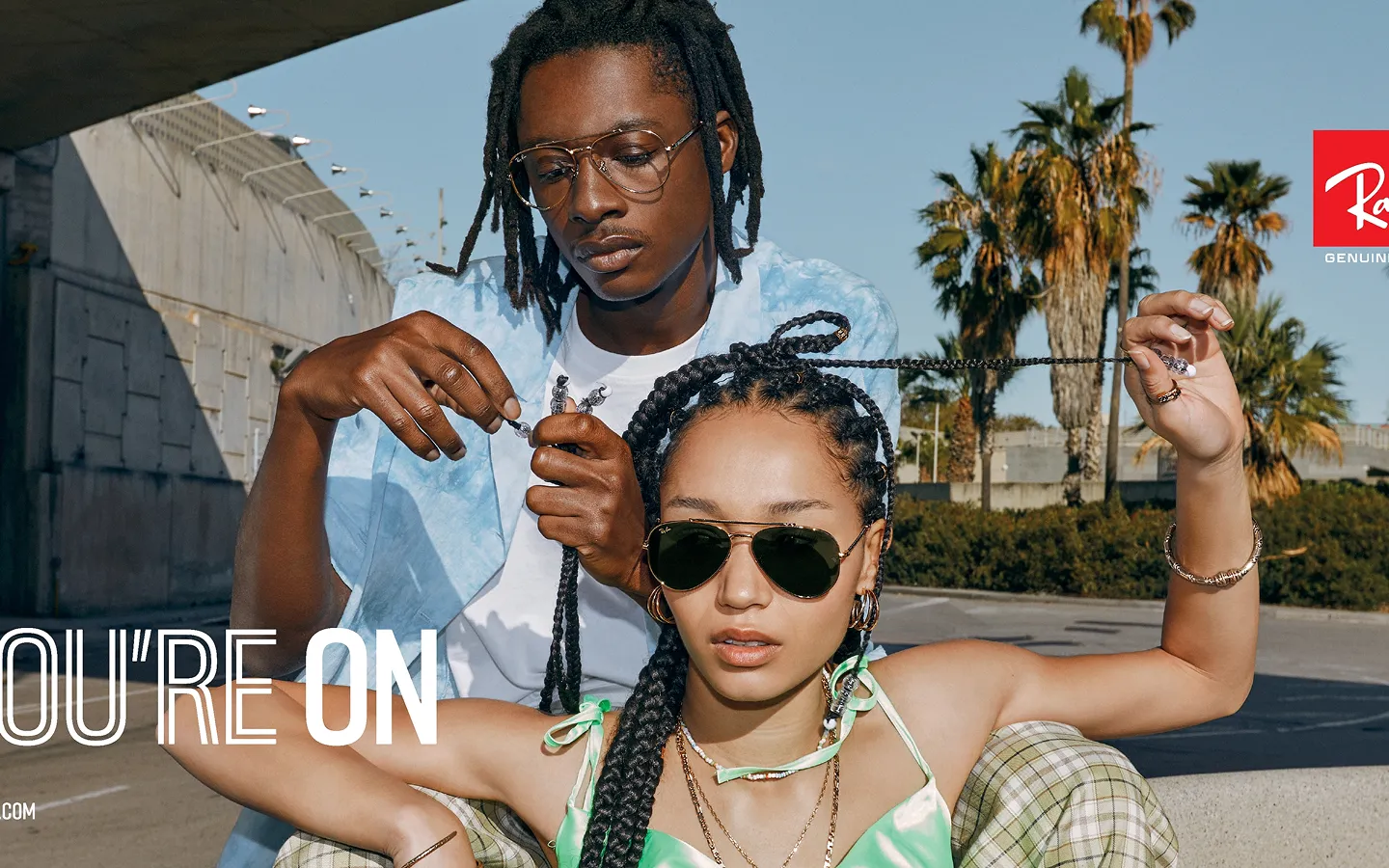 Man wearing gold-rimmed prescription glasses braiding a woman's hair; she wears aviator sunglasses and a green satin top outdoors with palm trees in the background.