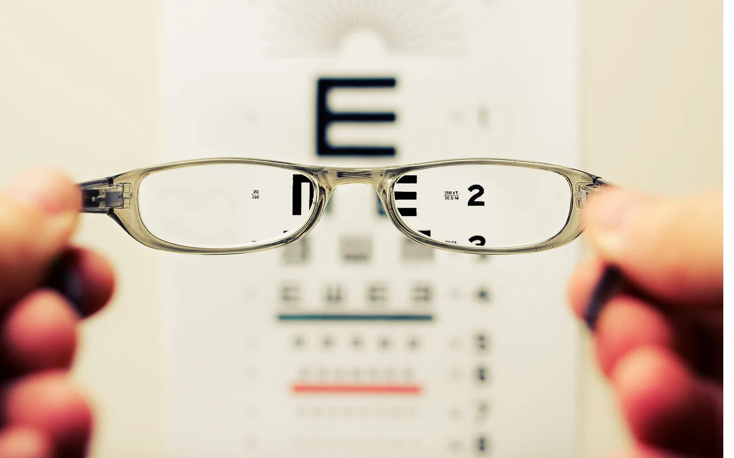 Close-up of hands holding eyeglasses focusing on a Snellen eye chart in the background.