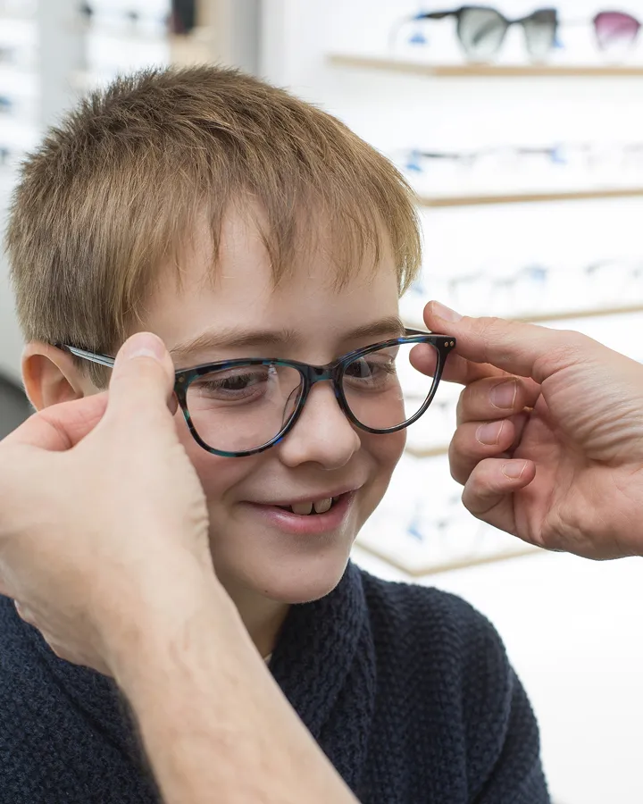 Smiling young boy trying on new eyeglasses with the help of an adult in a store.